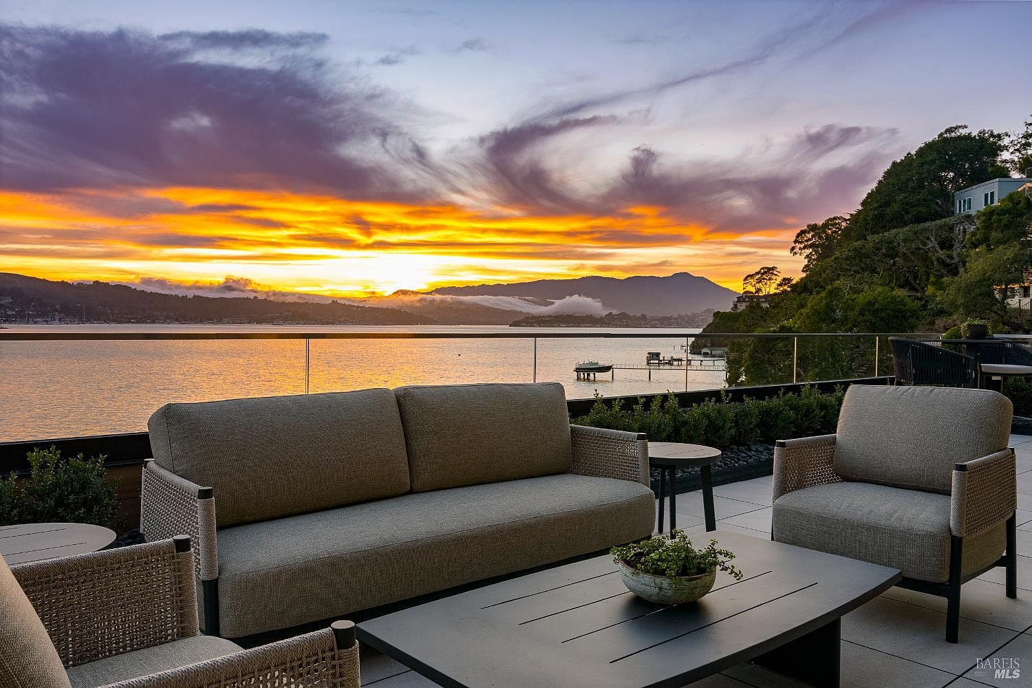 This expansive outdoor terrace offers a sophisticated seating area with a modern sofa, armchairs, and a low-profile coffee table, all arranged to maximize the panoramic water views. The space features a sleek glass railing that ensures an unobstructed perspective of the bay, distant mountains, and a vibrant sunset sky. The overall atmosphere is one of luxury and tranquility, perfect for evening relaxation or entertaining.