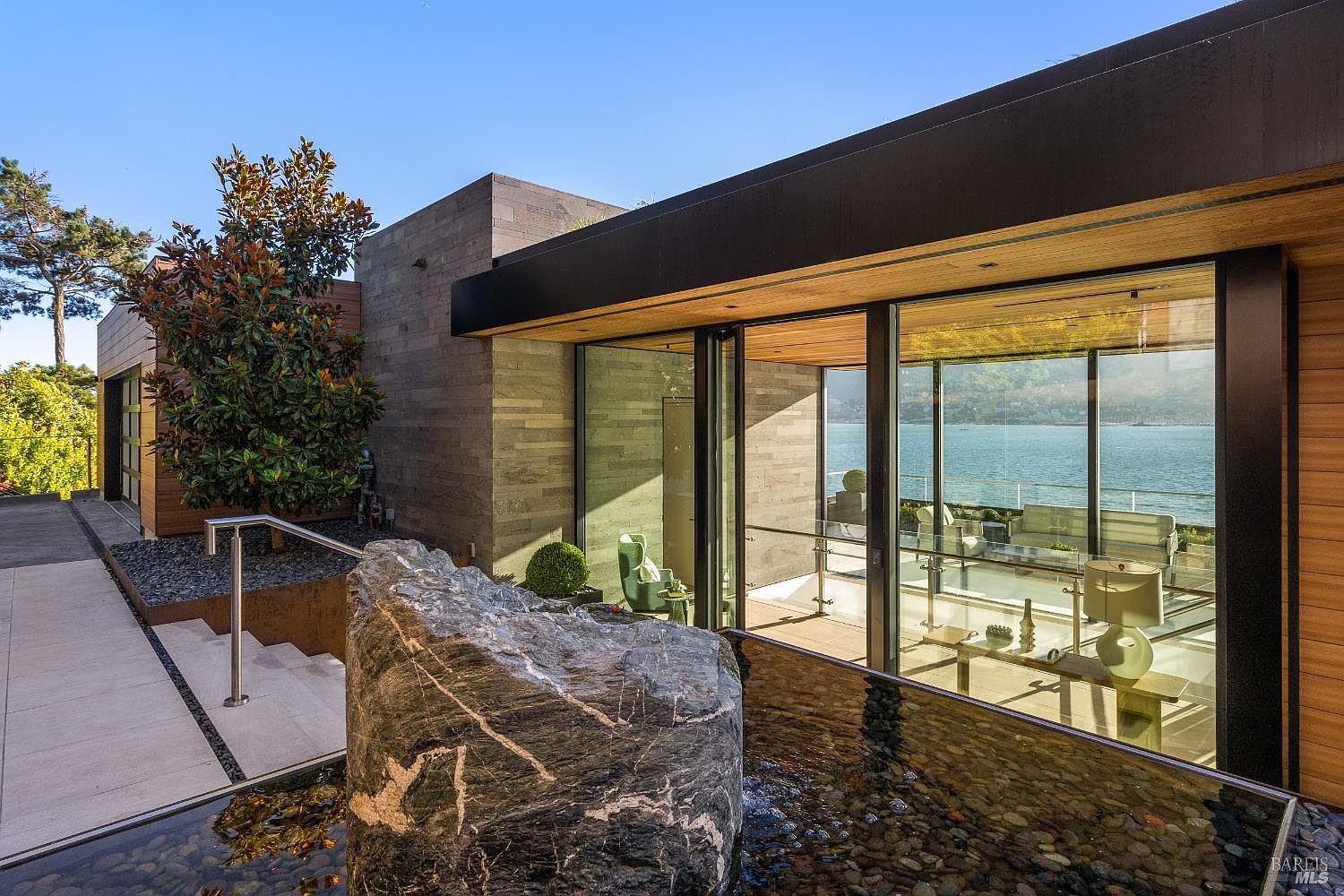 This modern entryway features a striking water feature with a large, textured stone centerpiece set in a shallow pool of dark pebbles. The home showcases contemporary architecture with clean lines, wood-paneled accents, and expansive glass walls that provide a clear view through the interior to the water beyond. A set of concrete steps with a sleek metal handrail leads toward the entrance, creating a sophisticated and welcoming transition into the residence.
