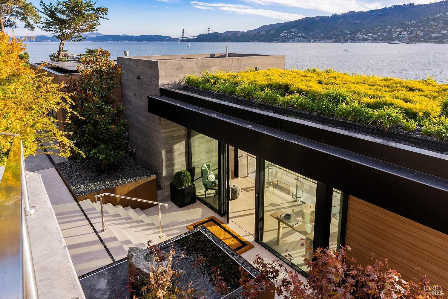 This high-angle exterior view captures a modern, multi-level residence featuring a green roof planted with vibrant yellow foliage. The home incorporates large glass sliding doors that open to a stone-paved patio, which includes a tranquil water feature and steps leading down to the lower level. In the background, the expansive bay and the iconic Golden Gate Bridge create a stunning, cinematic backdrop for this sophisticated waterfront property.