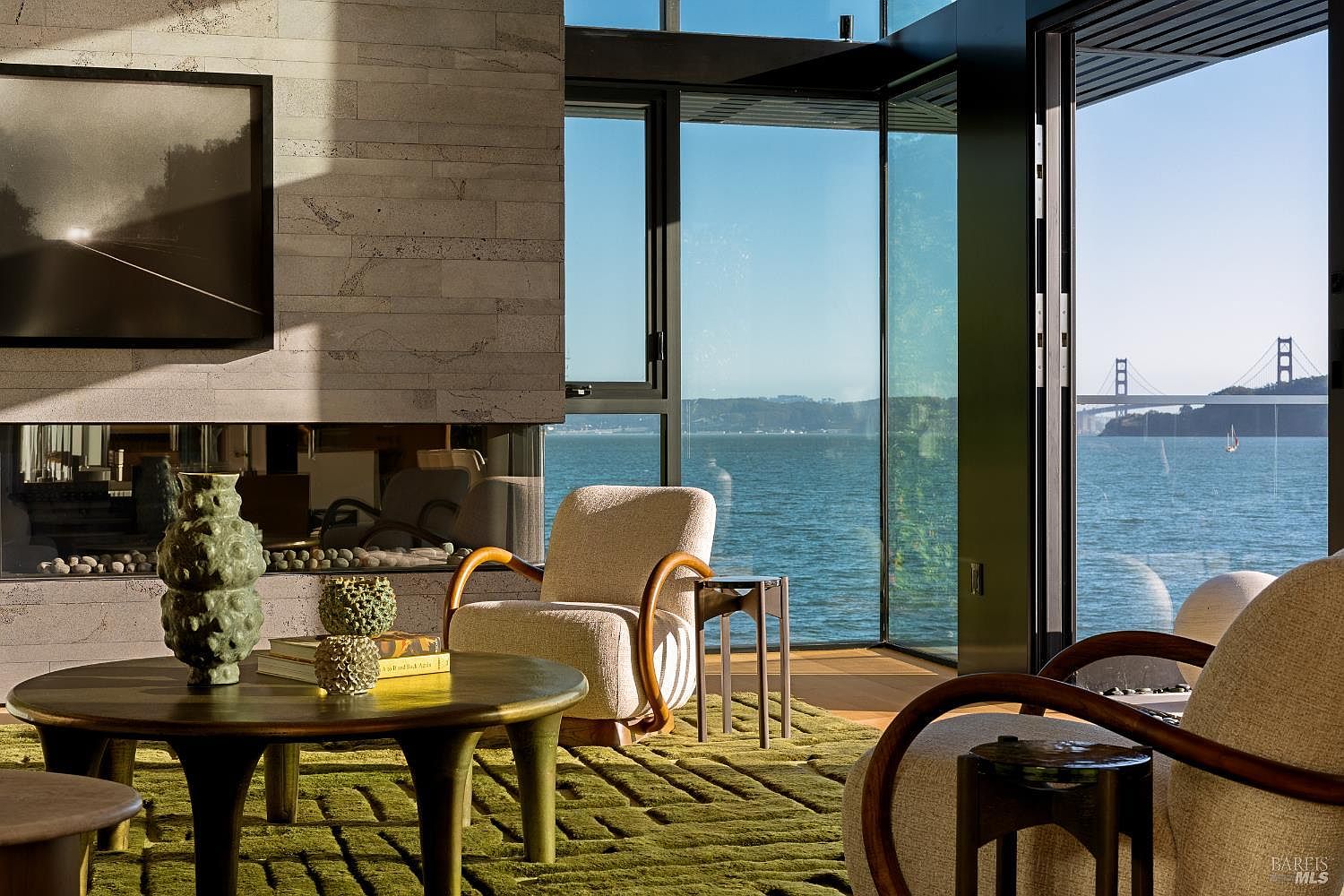 This sophisticated living space features a textured green area rug and mid-century modern style armchairs arranged around a circular wooden coffee table. A prominent stone-clad chimney breast anchors the room, while expansive glass walls provide an unobstructed, panoramic view of the bay and the Golden Gate Bridge. The perspective captures a serene, high-end atmosphere where the interior design seamlessly integrates with the iconic waterfront scenery.