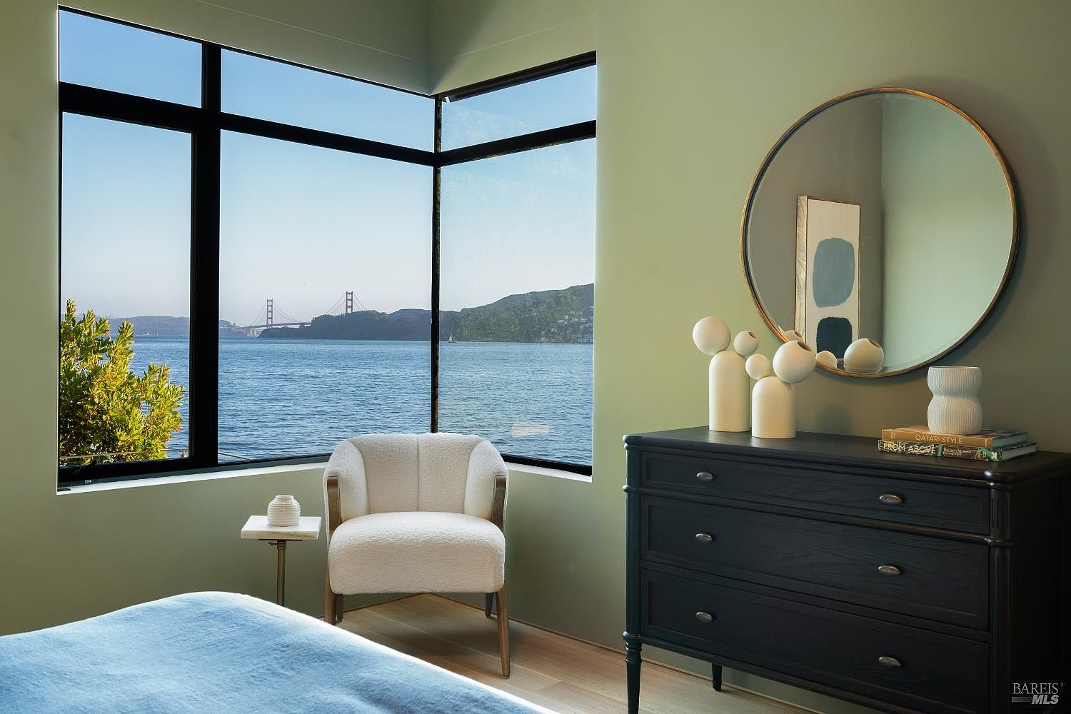 This serene bedroom features a corner window arrangement that frames a stunning, expansive view of the bay and the Golden Gate Bridge. The room is decorated in a calming palette of sage green walls, a textured cream-colored armchair, and a dark-toned dresser topped with minimalist decor and a circular mirror. The perspective captures a tranquil, high-end living space that seamlessly blends modern interior design with a dramatic natural backdrop.