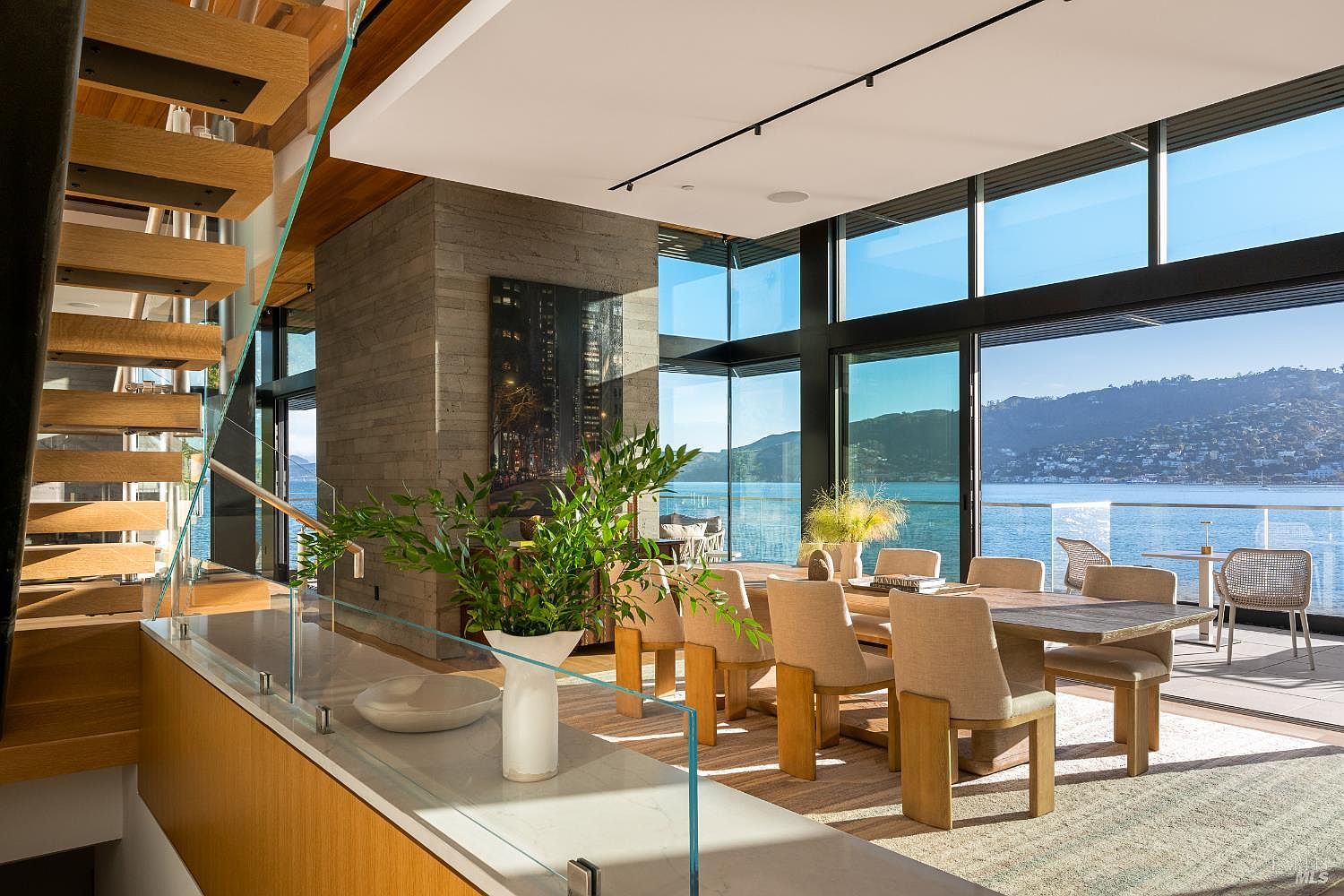 This sophisticated dining area features a large wooden table surrounded by upholstered chairs, set against a backdrop of expansive glass walls that offer panoramic water and mountain views. A prominent stone-clad chimney breast anchors the space, while a floating staircase with glass railings adds a modern architectural element to the foreground. The room is bathed in natural light, creating an airy and luxurious atmosphere that seamlessly connects the interior living space with the outdoor deck.