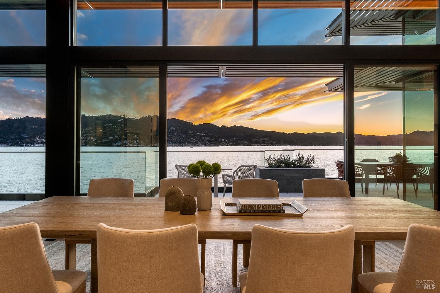 This elegant dining room features a large, light wood table surrounded by upholstered chairs, centered in front of expansive floor-to-ceiling glass walls that frame a stunning sunset over the water. The minimalist decor includes a simple vase with greenery and a decorative tray on the table, emphasizing the seamless connection between the indoor living space and the outdoor terrace. The perspective is captured from the dining area looking outward, highlighting the dramatic natural light and the serene, panoramic waterfront view.