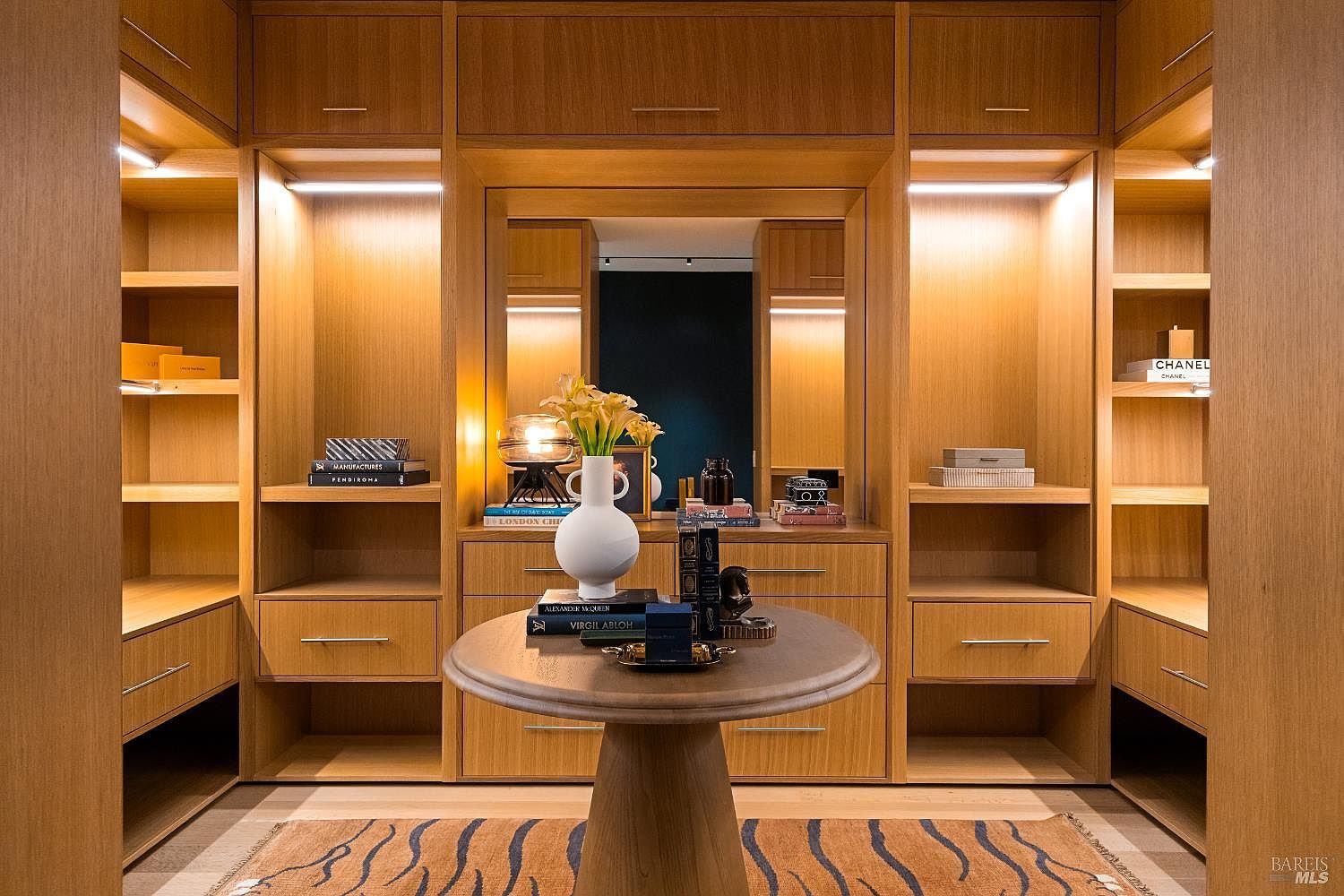 This sophisticated walk-in closet features custom light wood cabinetry with integrated LED lighting, providing a warm and organized display space. A central pedestal table serves as a focal point, topped with a decorative vase and designer books, while a patterned rug adds texture to the floor. The symmetrical layout, complete with a central mirror and ample shelving, creates a luxurious and boutique-like atmosphere.