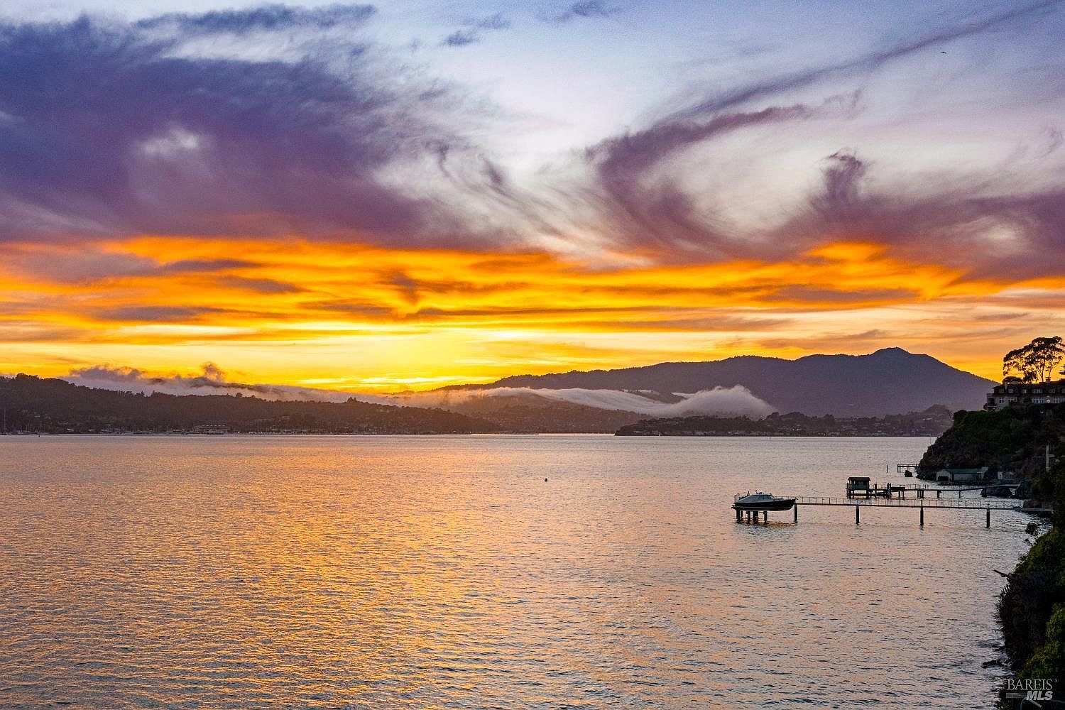 This scenic landscape view captures a vibrant sunset over a calm body of water, likely a bay or harbor. In the foreground, a private pier extends into the water with a small boat moored at the end, while distant mountains are partially shrouded in low-hanging clouds. The perspective is elevated, offering a panoramic outlook that emphasizes the tranquil and picturesque setting of the property's surroundings.
