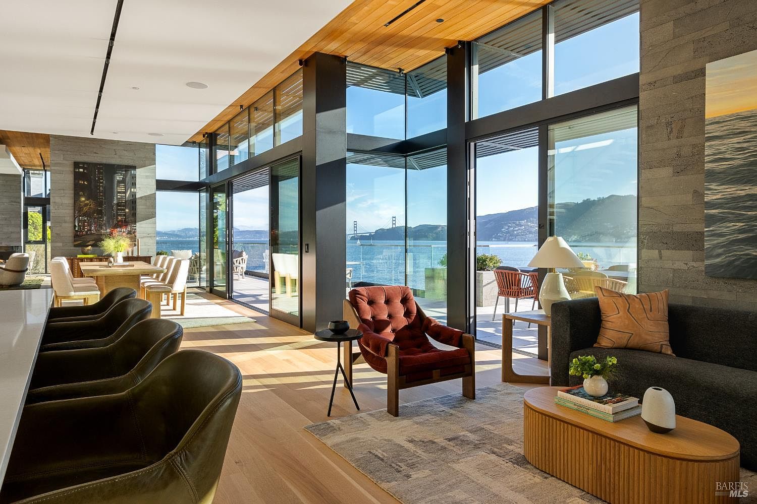 This sophisticated open-concept living space features a seamless transition to an outdoor deck through expansive glass walls that frame a stunning waterfront view, including the Golden Gate Bridge. The interior is defined by light wood flooring, a textured stone wall, and a warm, modern aesthetic with a plush velvet armchair and a contemporary sofa. The perspective captures the airy, light-filled atmosphere, highlighting the integration of indoor and outdoor living areas.