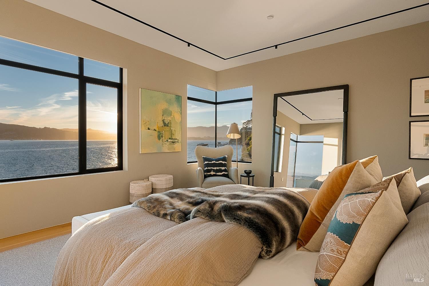 This serene primary bedroom features a plush bed dressed in layered neutral textiles and a faux-fur throw, positioned to take full advantage of the expansive water views. Large windows frame the scenic coastal landscape, while a tall, dark-framed mirror reflects the room's sophisticated, minimalist aesthetic. A cozy reading nook with an armchair and a floor lamp sits by the window, creating a tranquil retreat with a soft, warm color palette.