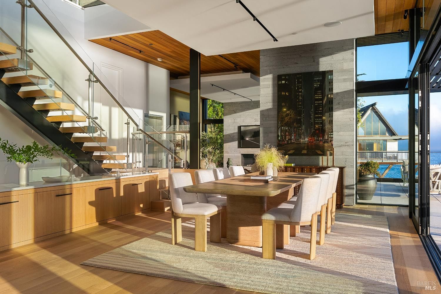 This sophisticated dining area features a large, light wood table surrounded by upholstered chairs, set against a backdrop of a stone-clad chimney breast. To the left, a floating staircase with glass railings adds a modern architectural element, while the right side showcases expansive glass walls that provide a seamless view of the water and outdoor deck. The space is bathed in warm, natural light, highlighting the clean lines and contemporary design of the home.