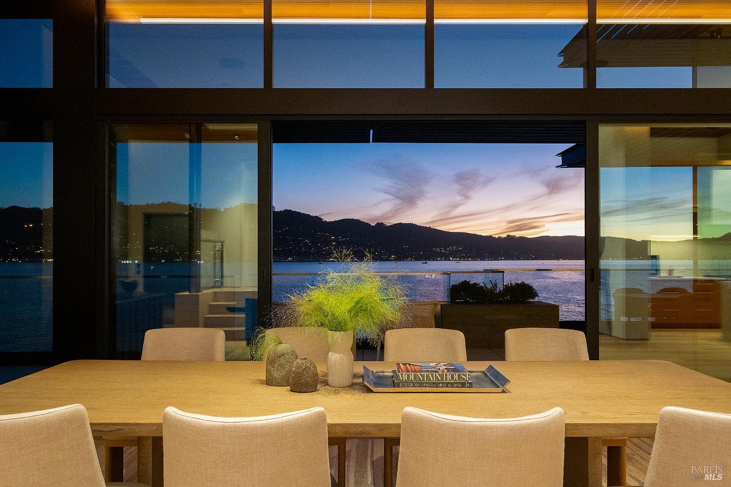 This elegant dining room features a long, light wood table surrounded by upholstered chairs, centered in front of expansive floor-to-ceiling glass walls that frame a stunning sunset view over the water and mountains. The minimalist decor includes a decorative tray with a book and a vase of greenery, emphasizing a serene and sophisticated atmosphere. The seamless transition between the interior space and the outdoor scenery creates a dramatic, cinematic backdrop perfect for refined entertaining.