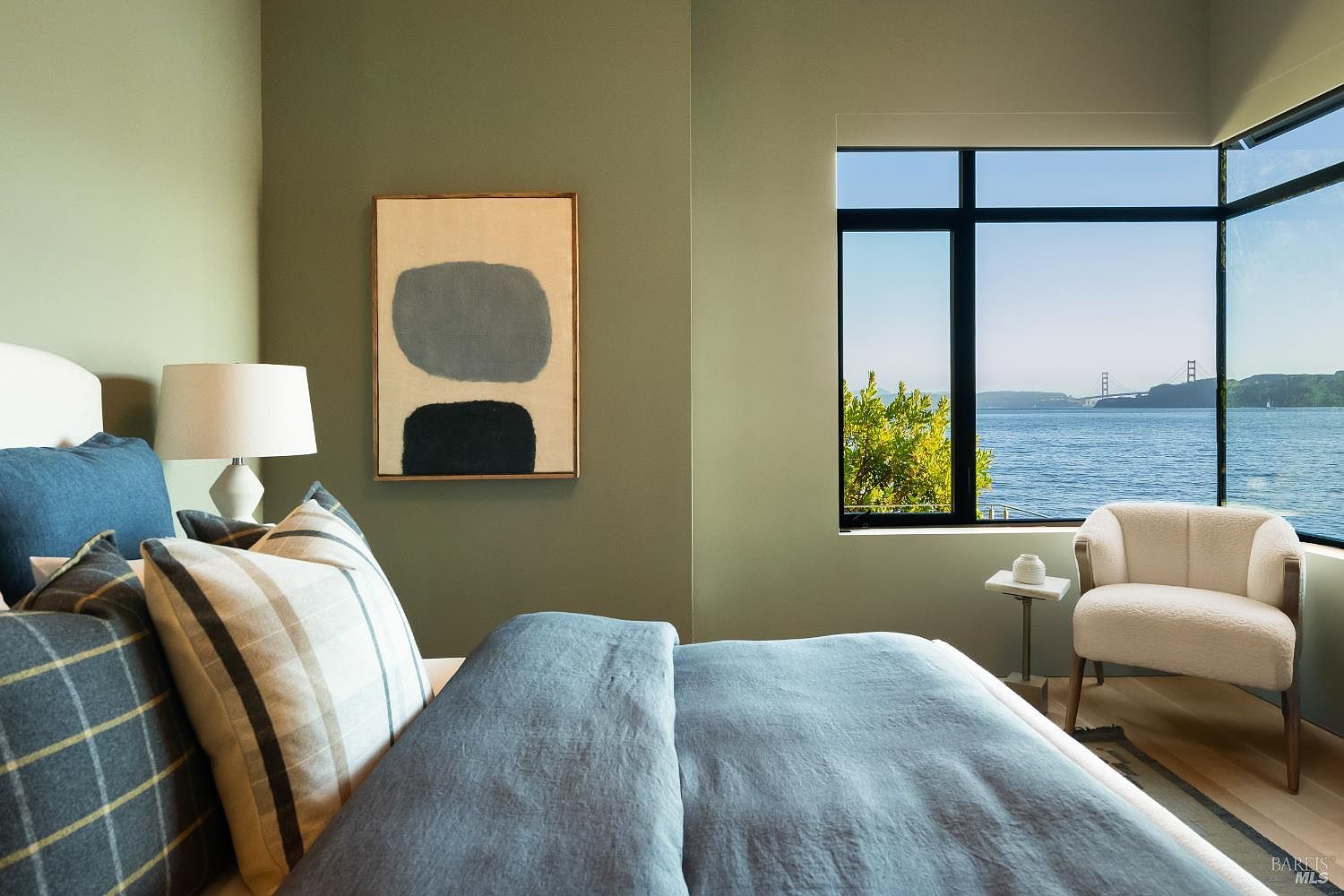 This serene bedroom features sage green walls and a plush bed dressed in blue and plaid linens, creating a calming atmosphere. A large window provides a stunning, unobstructed view of the bay and the Golden Gate Bridge, while a modern cream-colored armchair sits in the corner. The room is accented by a minimalist abstract painting and a bedside lamp, offering a sophisticated and tranquil retreat.
