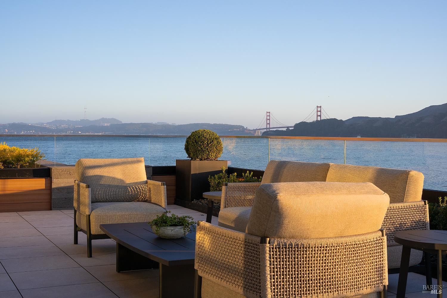This elevated outdoor terrace offers a sophisticated lounge area with comfortable, modern patio furniture arranged around a low coffee table. The space is defined by a sleek glass railing that provides an unobstructed, panoramic view of the bay and the iconic Golden Gate Bridge in the distance. The warm, golden-hour lighting enhances the inviting atmosphere, making it an ideal setting for relaxation or entertaining against a stunning coastal backdrop.