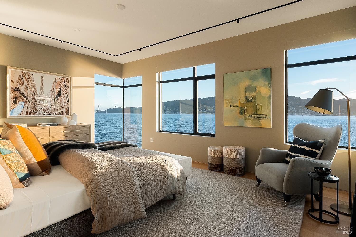 This primary bedroom features a contemporary design with a neutral color palette and expansive windows that provide panoramic water views. The room is furnished with a plush bed, a comfortable armchair, and a minimalist dresser, all set against a backdrop of soft, warm-toned walls. The space is bathed in natural light, creating a serene and luxurious atmosphere that emphasizes the connection to the waterfront setting.