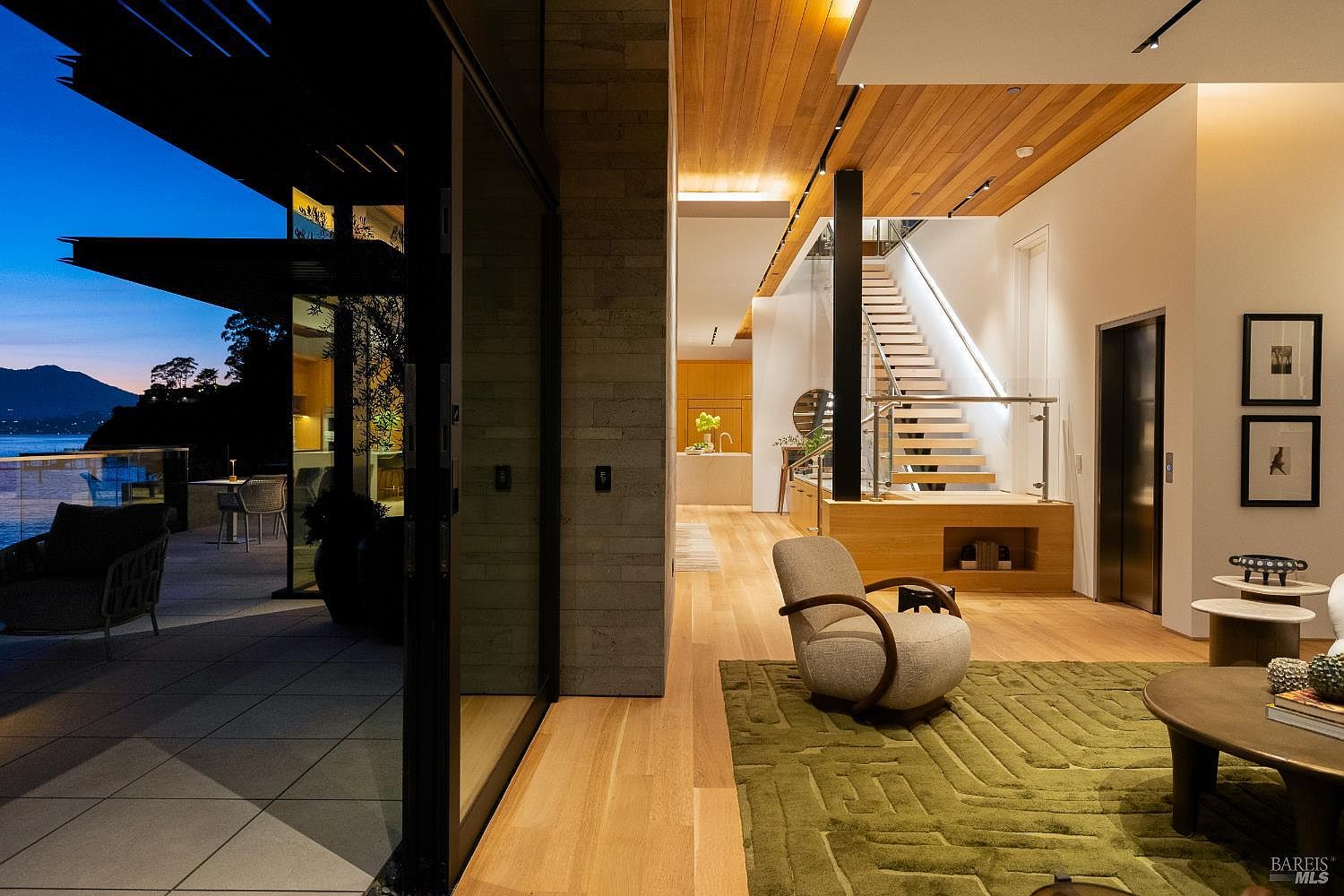 This sophisticated living space seamlessly blends indoor and outdoor living through expansive glass sliding doors that open onto a waterfront deck. The interior features warm light wood flooring, a textured green area rug, and a modern curved armchair, all set against a backdrop of a striking staircase with glass railings. The perspective captures a transition from the cozy, well-lit living area into a bright, open-concept kitchen and hallway, creating a sense of architectural depth and luxury.