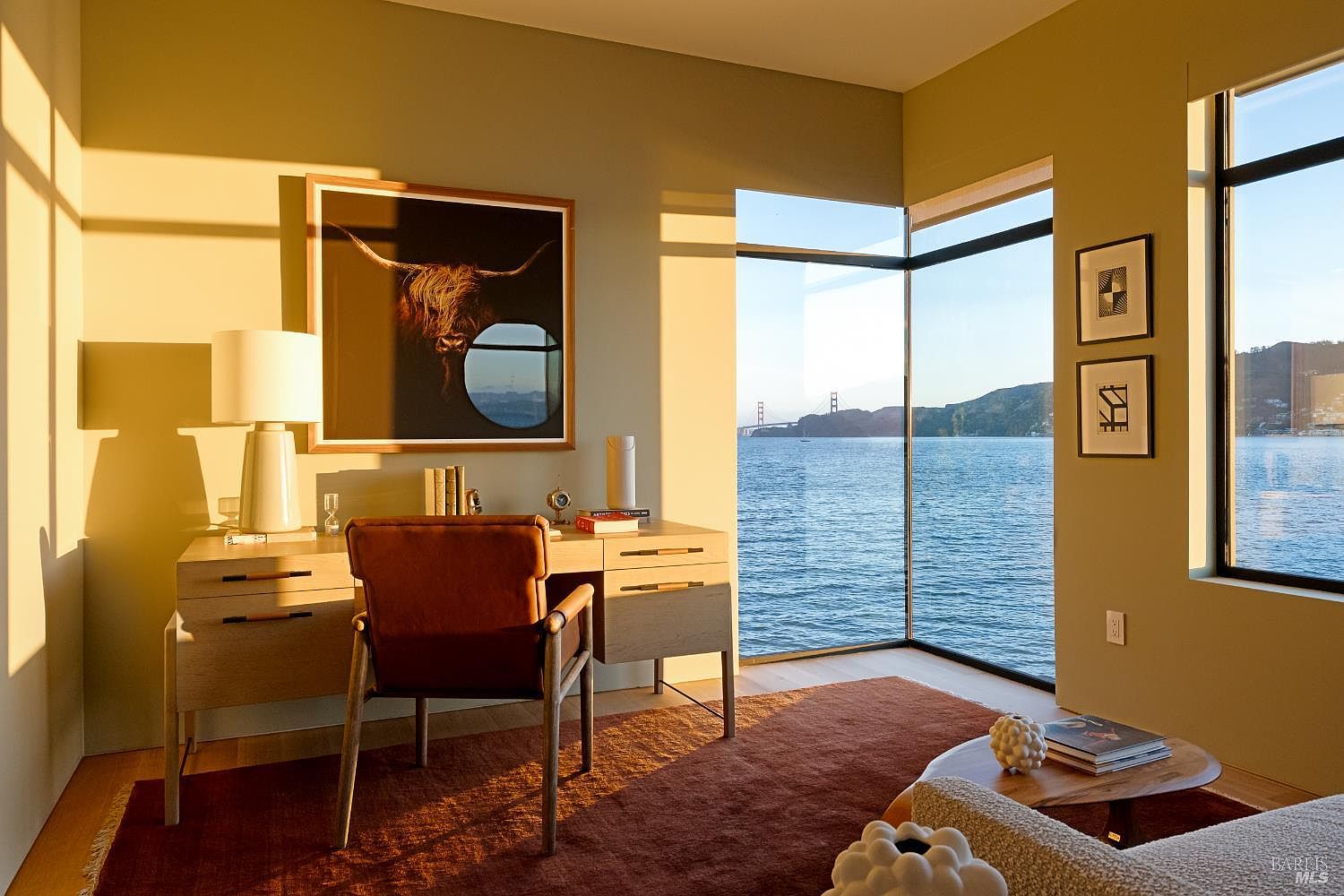 This sophisticated home office features a light wood desk paired with a leather chair, positioned to take full advantage of the stunning waterfront view through expansive corner windows. The room is bathed in the warm, golden light of sunset, highlighting a large framed artwork of a highland cow and a textured rug that anchors the space. The minimalist aesthetic and serene atmosphere create an ideal environment for focused work while overlooking the bay and distant bridge.