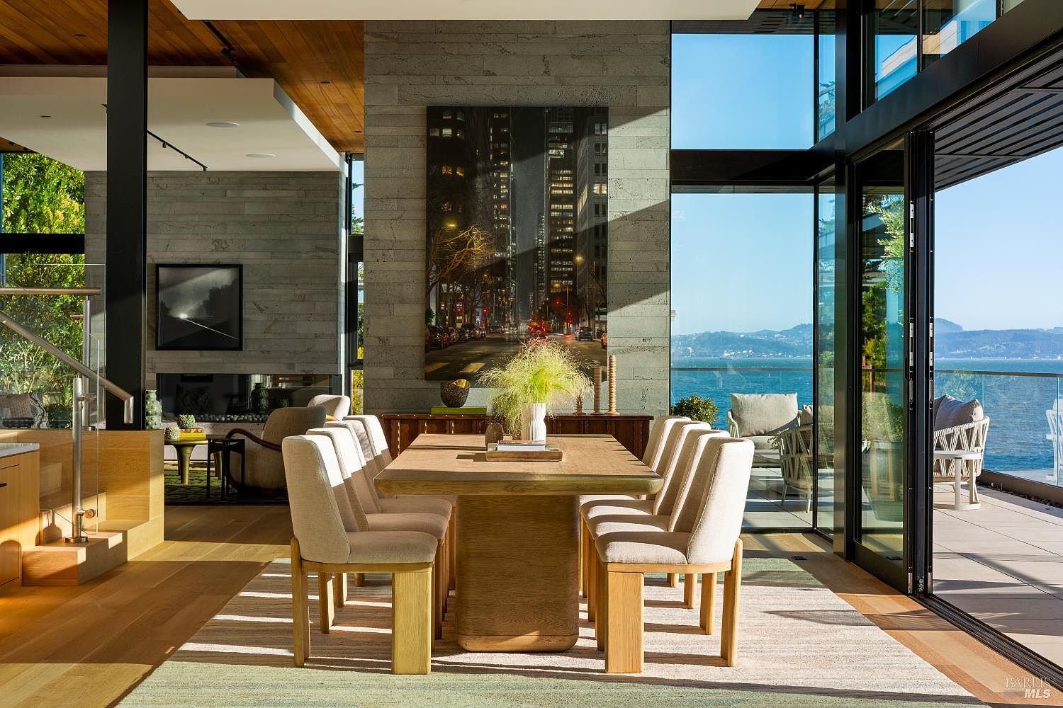 This sophisticated dining area features a light wood table surrounded by six upholstered chairs, set upon a neutral area rug. A prominent stone-clad chimney breast serves as a central architectural feature, adorned with a large vertical photograph of a city street. The space is bathed in natural light from expansive glass walls that provide a seamless transition to an outdoor deck and panoramic water views.