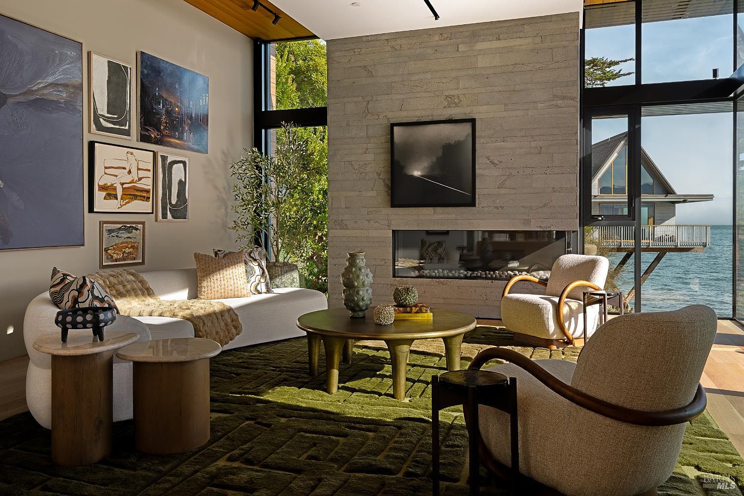 This sophisticated living room features a plush white sofa and two curved armchairs arranged around a circular coffee table on a deep green textured rug. A prominent stone-clad chimney breast houses a linear fireplace, serving as the room's focal point, while large windows provide expansive views of the water and a neighboring structure. The space is decorated with a gallery wall of framed art and a potted plant, creating a serene and luxurious atmosphere.
