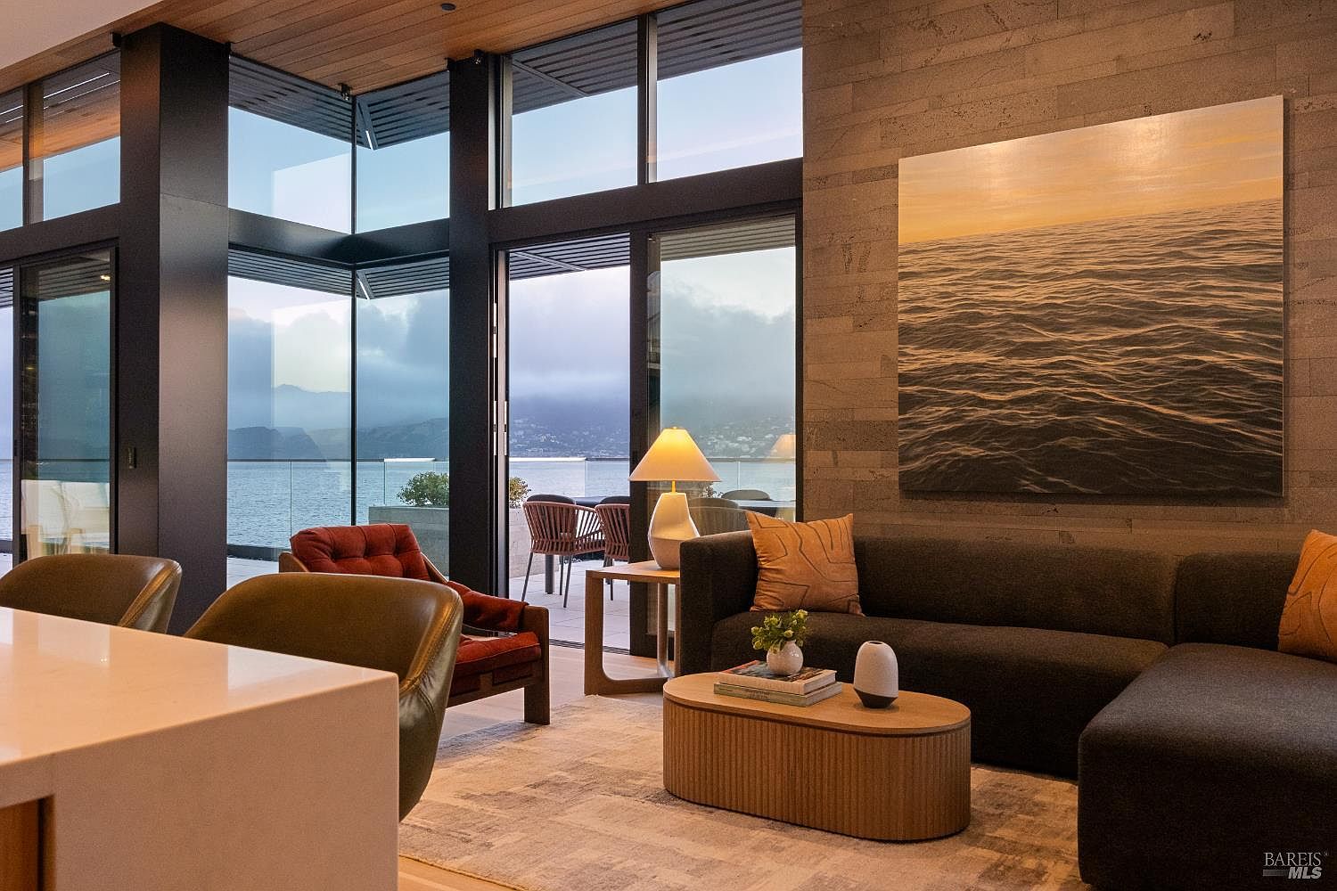This sophisticated living room features a dark, plush sectional sofa and a deep red accent chair, centered around an oval wooden coffee table. The space is defined by expansive floor-to-ceiling glass walls that frame a stunning panoramic view of the water and distant hills. A large, textured piece of art hangs on a prominent stone-clad wall, adding a dramatic focal point to the contemporary, open-concept design.