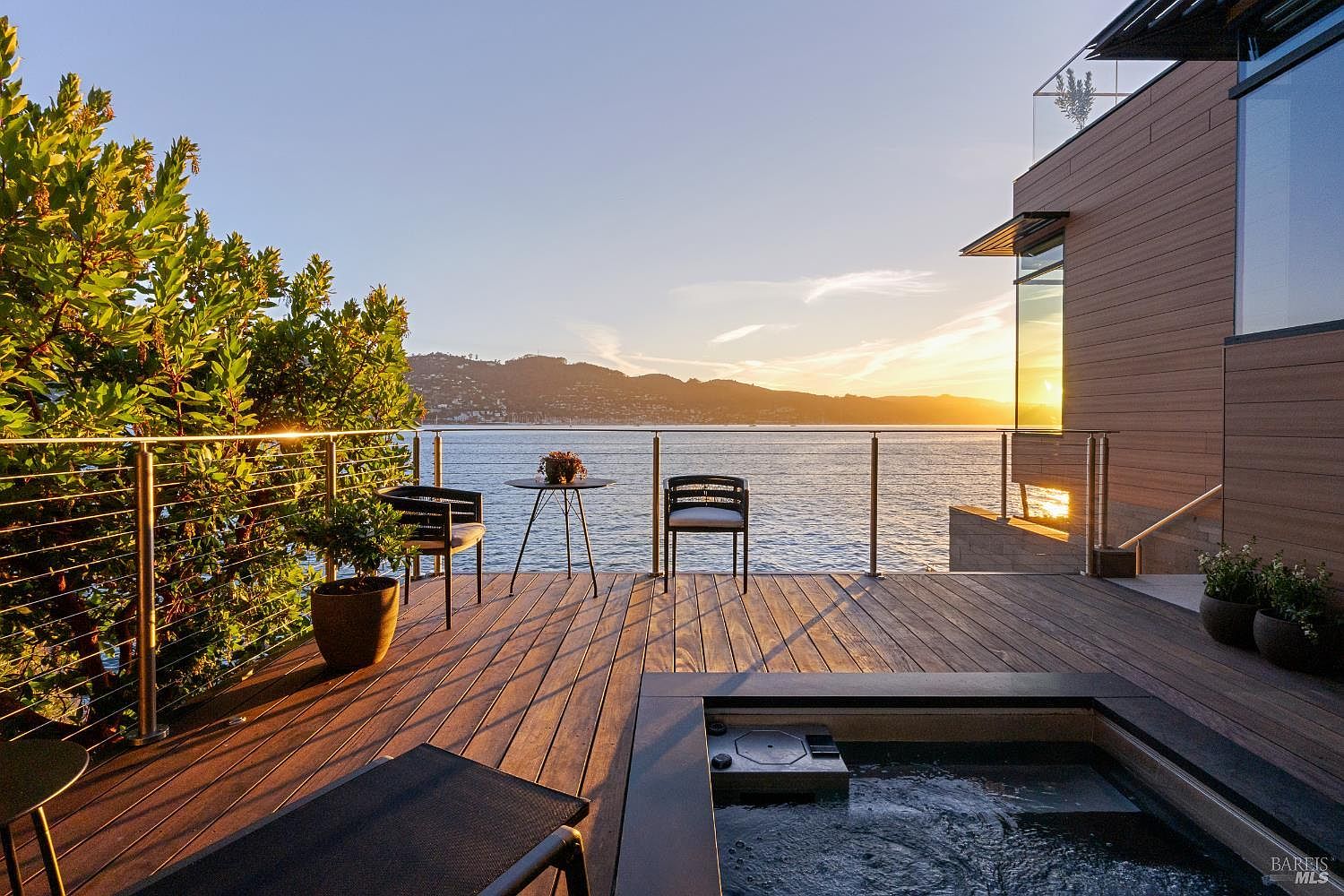 This stunning waterfront deck features a modern, minimalist design with warm-toned wood decking and a sleek metal railing that preserves the expansive bay views. The space is furnished with a small bistro set and a built-in spa, creating a serene atmosphere perfect for sunset relaxation. The perspective captures the seamless transition between the contemporary home's architecture and the natural beauty of the water and distant hills.