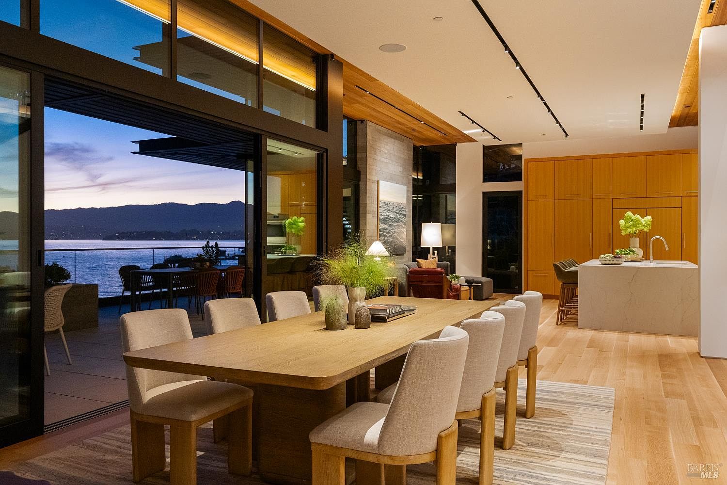 This sophisticated open-concept dining area features a large light wood table surrounded by upholstered chairs, seamlessly transitioning to an outdoor deck through expansive glass sliding doors. The space is anchored by a kitchen with light wood cabinetry and a stone-topped island, while a stone-clad wall adds texture to the living area beyond. The room is bathed in the soft, ambient light of dusk, highlighting the panoramic water and mountain views visible through the glass.
