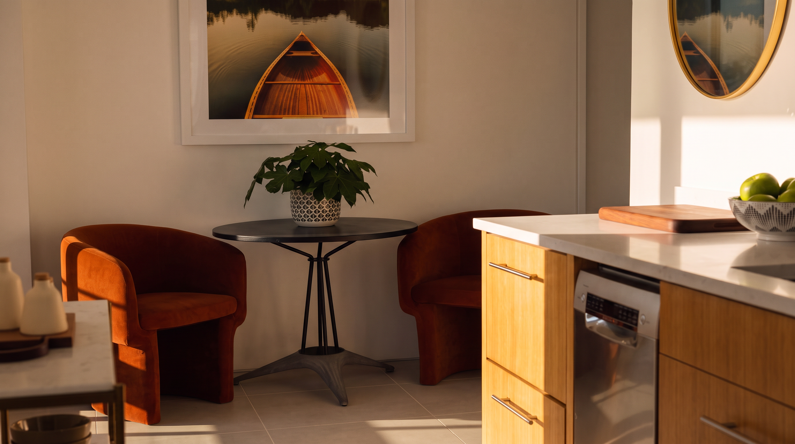 This bright, modern kitchen area features light wood cabinetry and a clean stone countertop that extends into a cozy breakfast nook. The nook is furnished with two vibrant orange chairs and a small round table, positioned beneath a framed landscape photograph. The perspective captures the seamless transition between the functional kitchen workspace and the intimate dining corner, highlighted by natural light streaming in from the side.