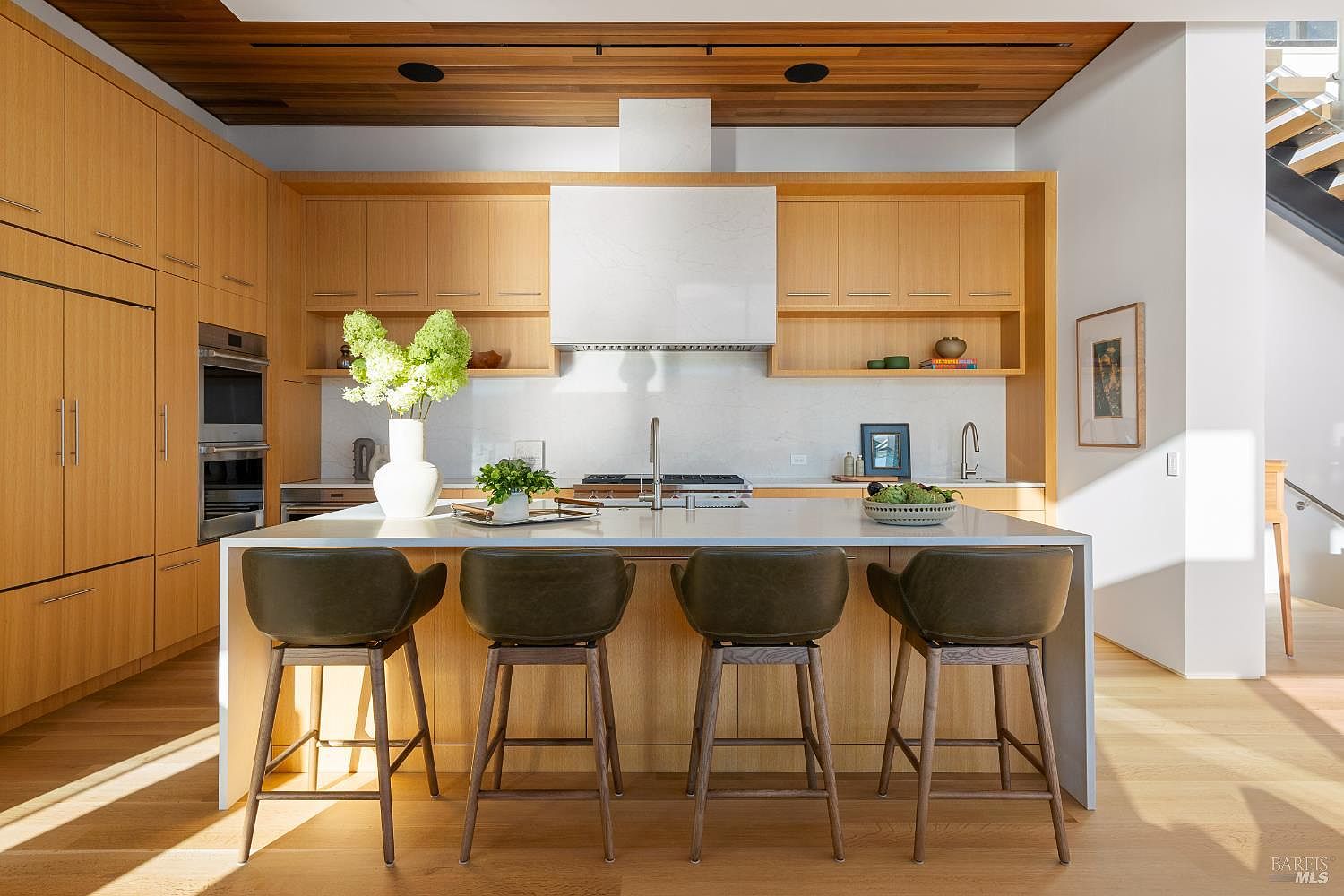 This modern kitchen features light wood cabinetry and a large central island topped with a white stone surface, providing ample seating with four dark-toned stools. The space is defined by a clean, minimalist aesthetic, highlighted by a white stone backsplash and a matching range hood. A warm, slatted wood ceiling adds texture and depth to the room, while natural light streams in from the right, creating an inviting and airy atmosphere.