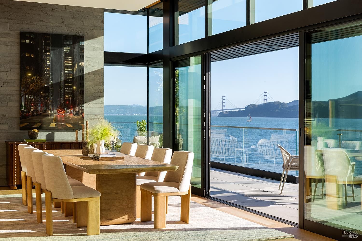 This sophisticated dining room features a large, light wood table surrounded by upholstered chairs, set against a backdrop of expansive floor-to-ceiling glass walls that frame a stunning view of the Golden Gate Bridge. To the left, a textured stone wall displays a large, vibrant cityscape artwork, while the open-plan design seamlessly connects the interior space to a private outdoor balcony. The room is bathed in natural light, creating an airy and luxurious atmosphere that highlights the seamless integration of indoor and outdoor living.
