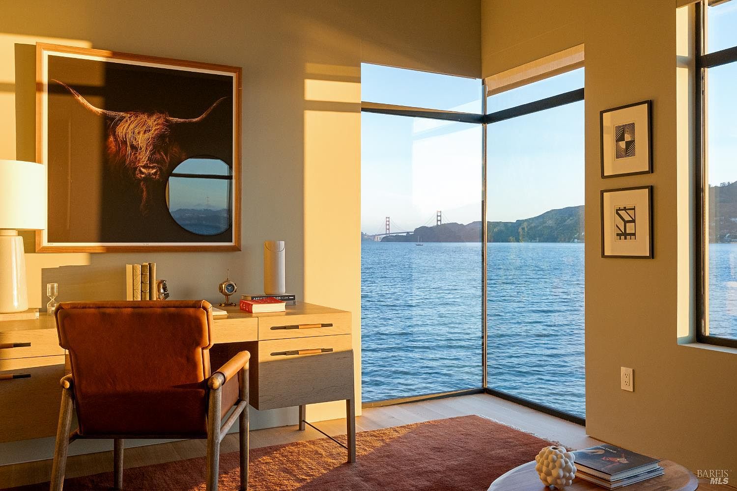 This sophisticated home office features a warm, minimalist aesthetic with a light wood desk and a leather chair positioned to take advantage of a stunning corner view. Large, floor-to-ceiling windows frame a panoramic vista of the water and the Golden Gate Bridge, bathing the room in golden hour light. The space is accented by a large, striking piece of wall art featuring a highland cow and two smaller framed prints, creating a serene and professional atmosphere.