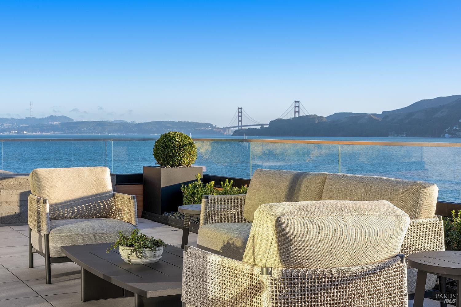 This expansive outdoor terrace offers a sophisticated lounge area furnished with contemporary, textured-weave seating and a low-profile coffee table. The space is defined by a sleek glass railing that provides an unobstructed, panoramic view of the Golden Gate Bridge and the surrounding bay. The setting is bright and airy, emphasizing a luxurious lifestyle with a seamless connection between comfortable outdoor living and iconic coastal scenery.