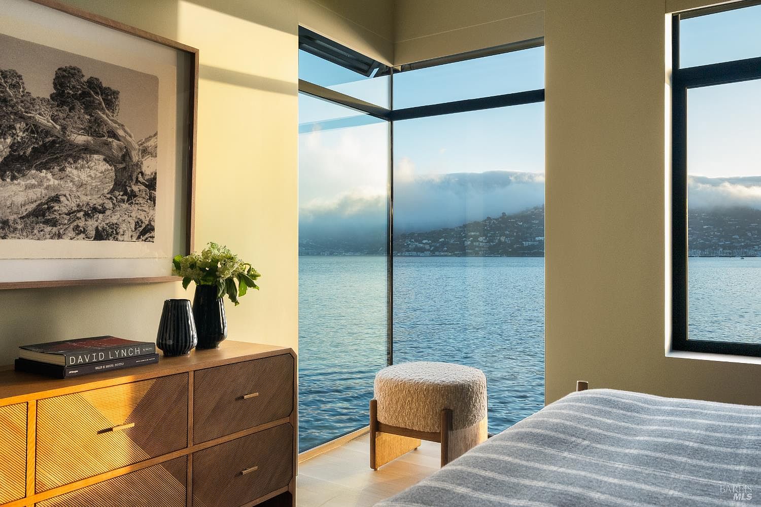 This serene bedroom features a corner window arrangement that provides expansive, unobstructed views of the water and distant hills. A textured wooden dresser sits to the left, topped with a vase of flowers and a book, while a plush, upholstered ottoman is positioned near the glass. The room is bathed in warm, natural light, creating a tranquil and sophisticated atmosphere.