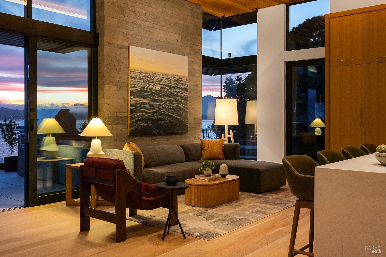 This sophisticated living room features a contemporary sectional sofa, a unique wooden coffee table, and a dark-toned accent chair, all arranged on a textured area rug. The space is defined by a prominent stone-clad wall and expansive glass walls that provide panoramic views of the water and sunset. Warm ambient lighting from multiple table lamps creates an inviting atmosphere, while the open-concept layout seamlessly connects to a kitchen area in the foreground.