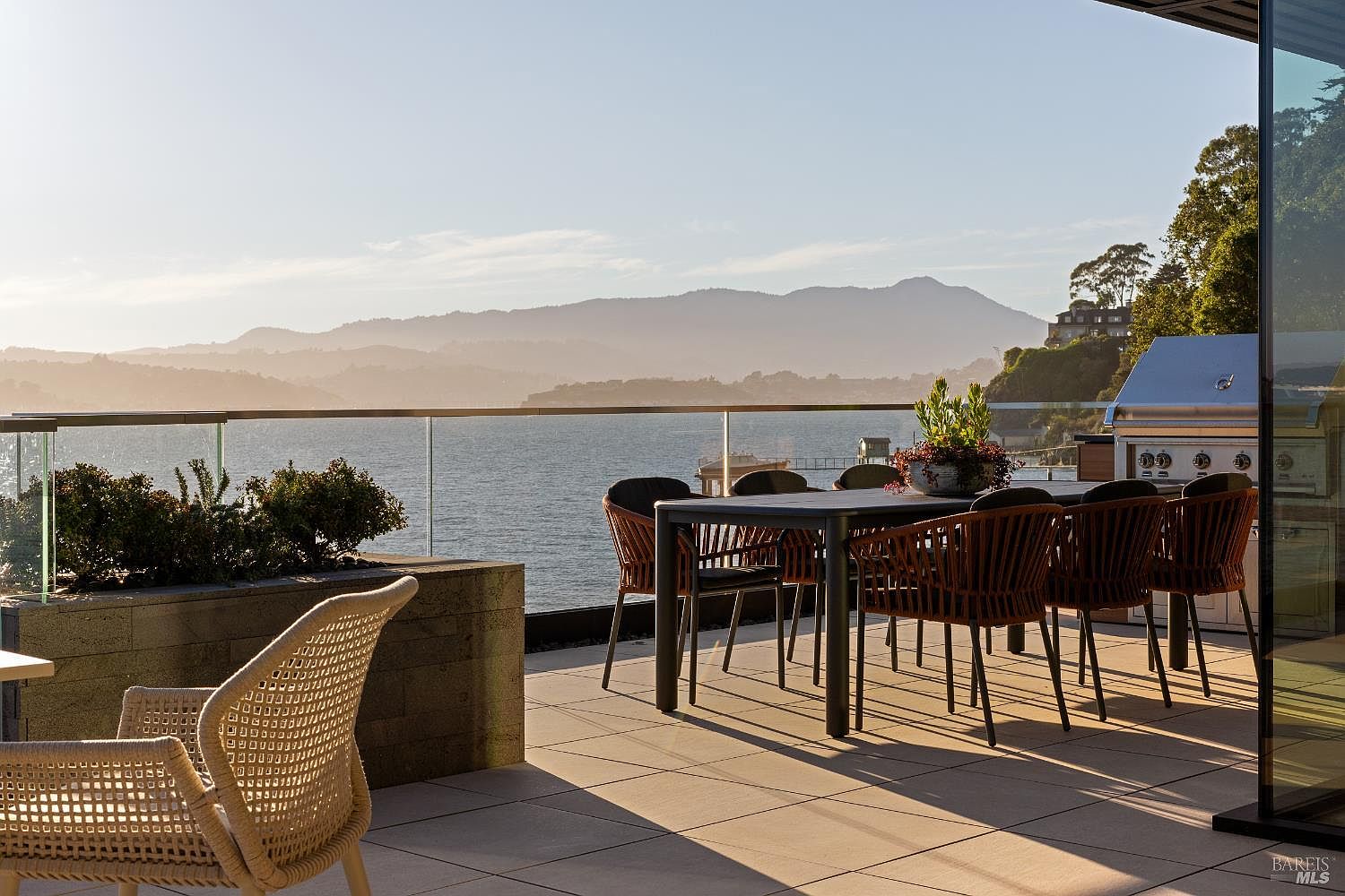 This expansive outdoor terrace offers a sophisticated dining area with a dark-toned table and woven chairs, set against a stunning panoramic view of the bay and distant mountains. A glass railing provides an unobstructed perspective, while a built-in grill station and a stone planter add functional elegance to the space. The scene is bathed in the warm, golden light of late afternoon, creating a serene and luxurious atmosphere perfect for entertaining.
