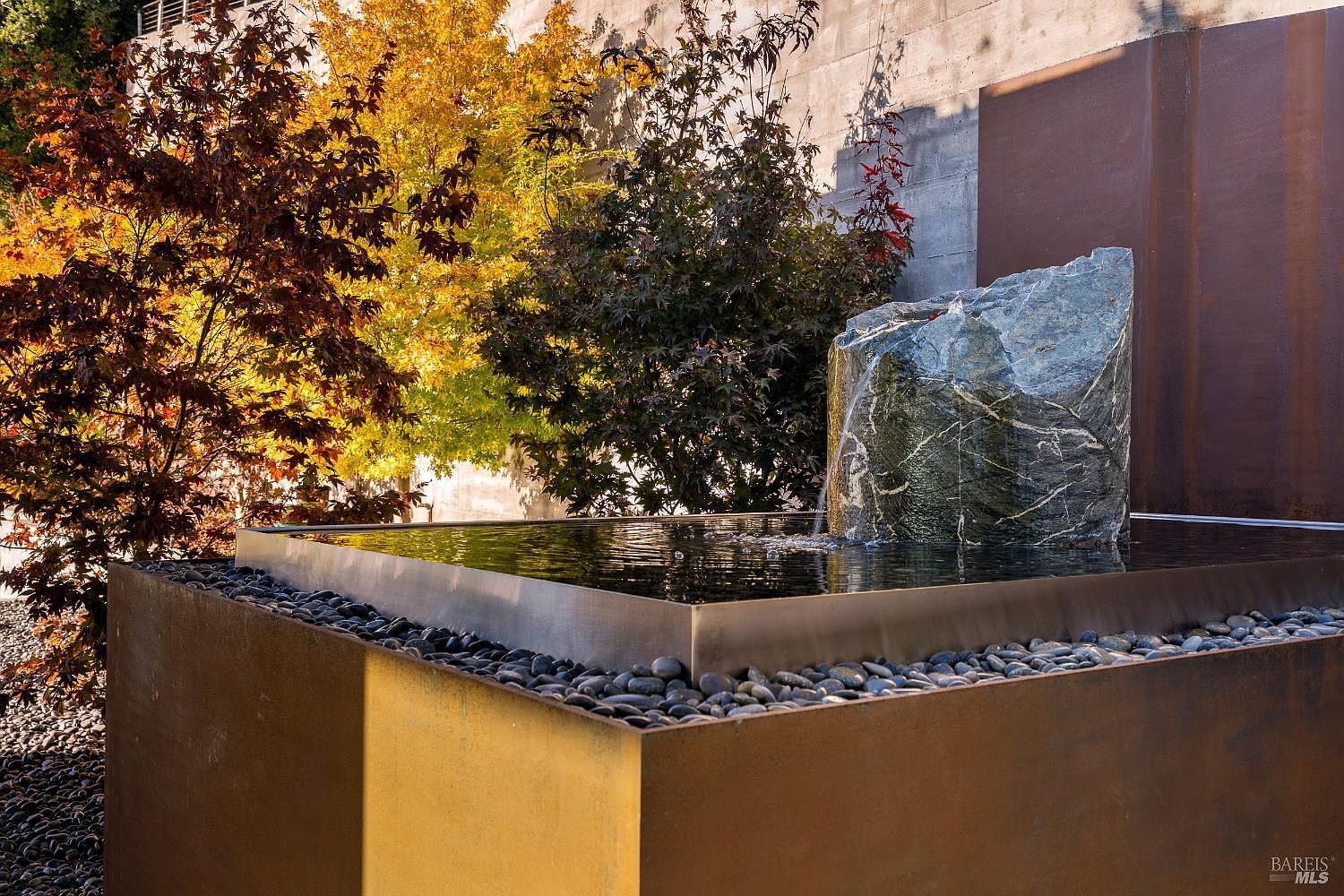 This image captures a modern, sculptural water feature set within a landscaped garden. A large, textured stone monolith serves as the centerpiece, with water cascading into a sleek, rectangular metal basin filled with smooth river stones. The scene is framed by vibrant autumn foliage in shades of gold and deep red, set against a minimalist concrete and metal wall backdrop.