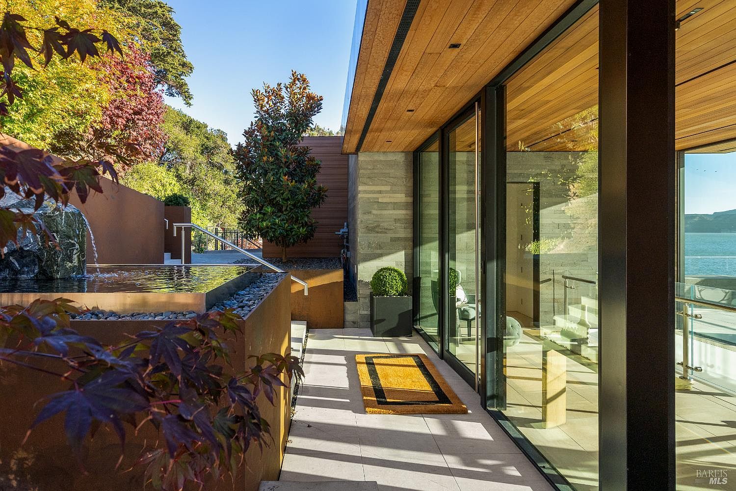 This modern outdoor patio space features a sleek water feature with a stone fountain and a minimalist metal basin. The area is defined by large glass sliding doors that provide a seamless transition to the interior, while a wood-paneled ceiling overhang adds warmth to the contemporary design. A yellow rug sits on the light-colored stone pavers, and the background offers a scenic view of the water and surrounding landscape.