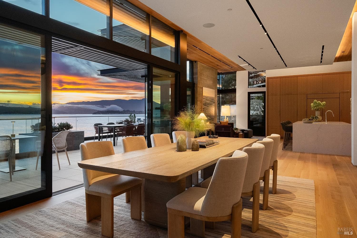 This sophisticated dining area features a large light wood table surrounded by upholstered chairs, set against a backdrop of expansive glass walls that open to a scenic waterfront deck. The open-concept space flows seamlessly into a modern kitchen with a central island and light wood cabinetry, all illuminated by recessed lighting and warm ambient glow. The overall aesthetic is contemporary and luxurious, emphasizing the connection between the indoor living space and the dramatic sunset views over the water.