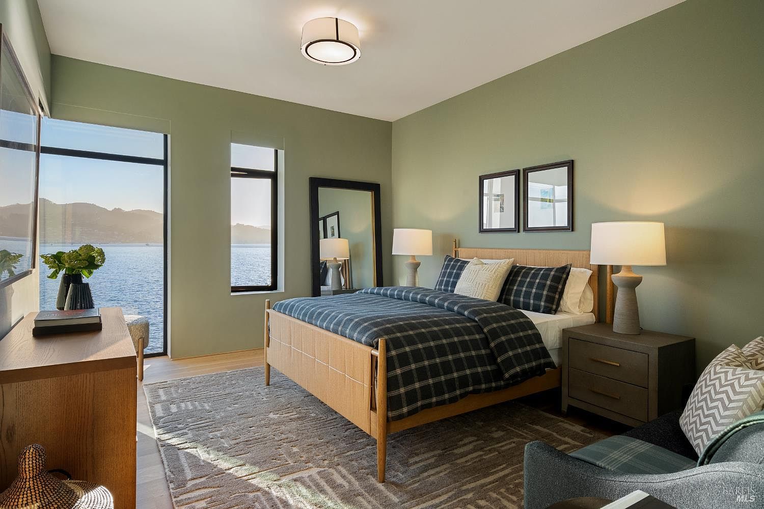 This serene primary bedroom features a light wood bed frame dressed in a dark plaid comforter, complemented by matching nightstands and table lamps. A large mirror leans against the sage green wall, while expansive windows provide a stunning, unobstructed view of the water and distant hills. The room is anchored by a textured area rug, creating a calm and sophisticated atmosphere.