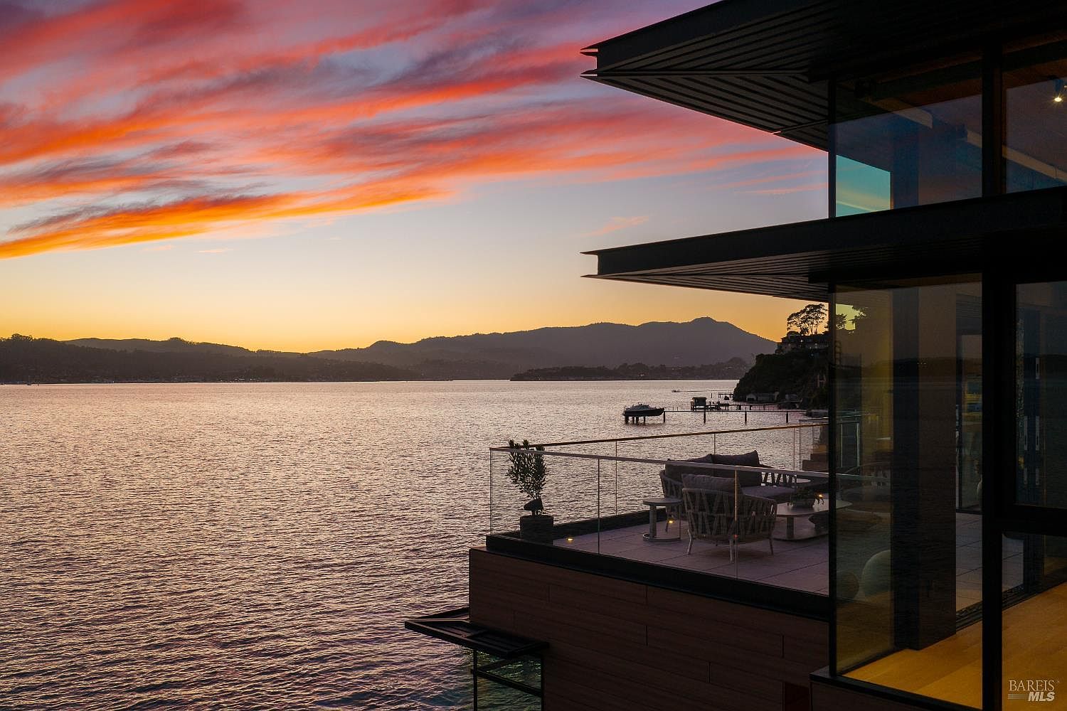 This stunning contemporary home features a spacious balcony overlooking a serene bay at sunset. The architectural design emphasizes indoor-outdoor living with expansive glass walls and sleek, modern lines. The balcony is furnished with comfortable seating, creating an ideal space to enjoy the vibrant orange and purple hues of the evening sky over the water.