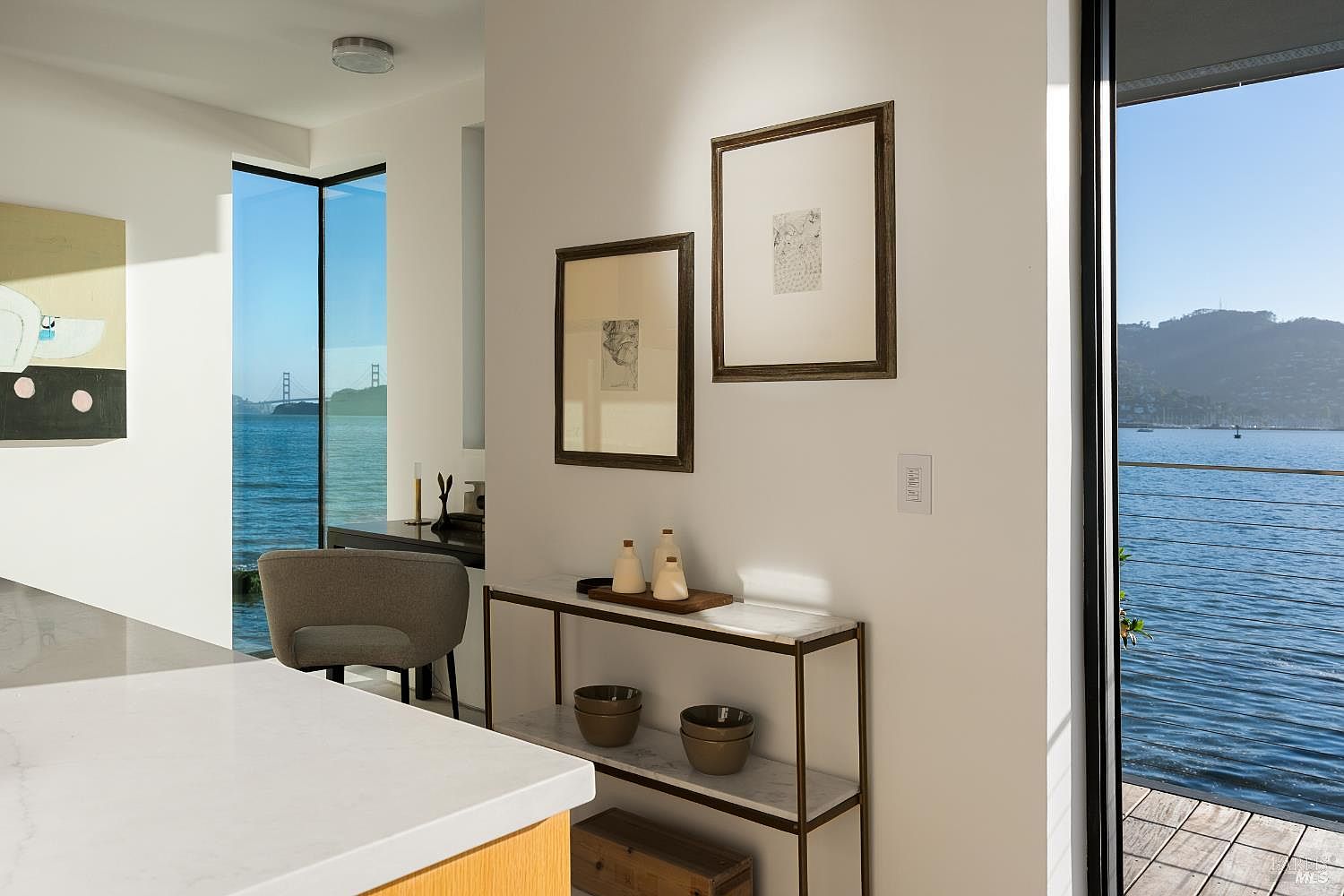 This interior view captures a bright, modern kitchen area featuring a light-toned stone countertop island in the foreground and a sleek console table against a white wall. The space is accented by framed artwork, minimalist decor, and a gray upholstered chair, all framed by expansive glass walls that offer a stunning, unobstructed view of the bay and distant hills. The composition emphasizes a seamless transition between the refined indoor living space and the natural beauty of the waterfront setting.