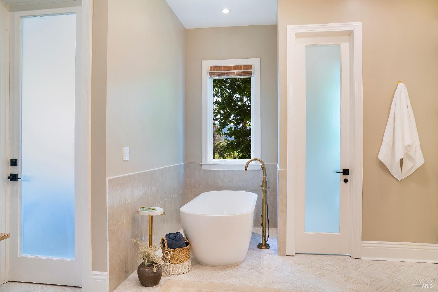 This elegant primary bathroom features a sleek, freestanding white soaking tub as its centerpiece, complemented by a vintage-style brass floor-mounted faucet. The space is finished with light beige wall paint, a herringbone-patterned tile floor, and two frosted glass doors that provide privacy while maintaining a bright, airy atmosphere. A small side table, a woven basket with towels, and a potted plant add a touch of warmth and texture to the clean, modern design.