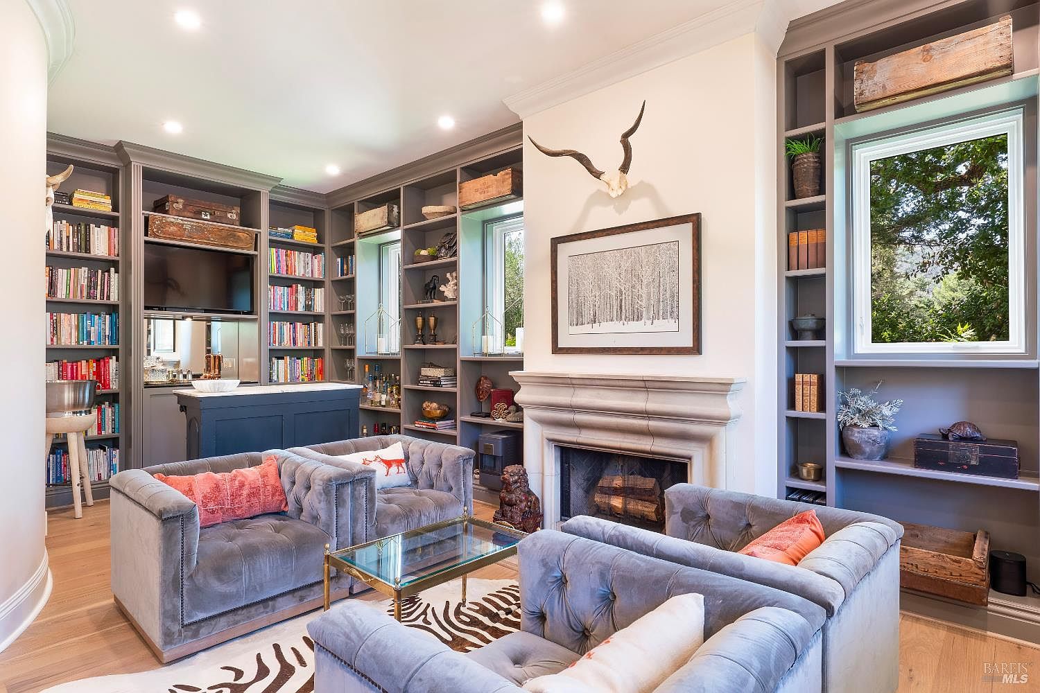 This sophisticated living room features a symmetrical layout with two plush, grey tufted velvet sofas facing a central fireplace adorned with a decorative animal skull. Built-in floor-to-ceiling grey shelving units flank the fireplace and provide ample storage for books and decor, while a glass-topped coffee table sits atop a stylish zebra-print rug. The room exudes a refined, cozy atmosphere, enhanced by warm recessed lighting and a view of lush greenery through the side window.