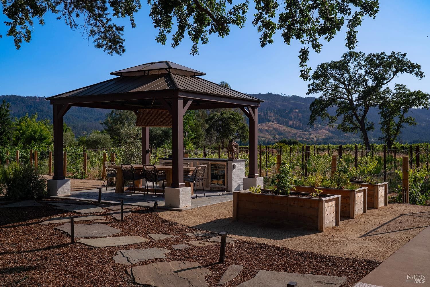 This picturesque outdoor living space features a covered gazebo with a dining area and built-in outdoor kitchen, set against a backdrop of rolling hills and a vineyard. In the foreground, raised wooden garden beds and a stone pathway through mulch create a charming, rustic aesthetic. The scene is bathed in warm, natural light, emphasizing the serene and expansive nature of the property.