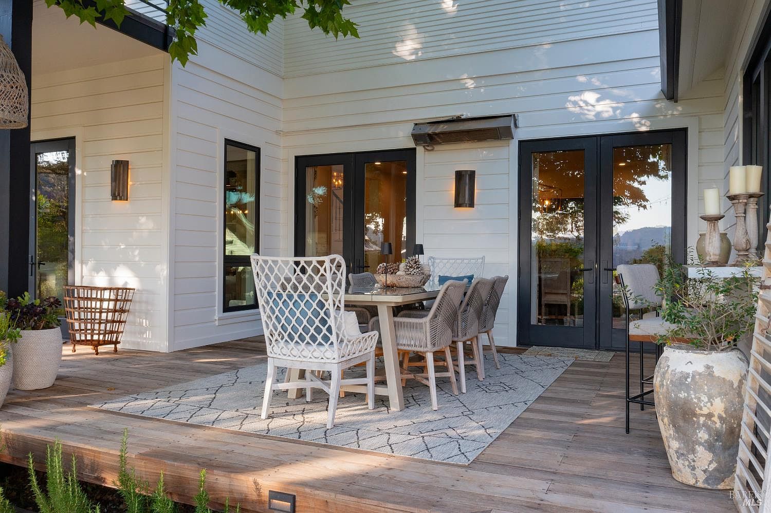This inviting outdoor deck features a modern farmhouse aesthetic with white horizontal siding, dark-framed French doors, and natural wood decking. A stylish dining set with woven chairs sits atop a patterned area rug, creating a perfect space for al fresco dining. The scene is framed by lush greenery and warm lighting, offering a serene and sophisticated atmosphere for outdoor living.