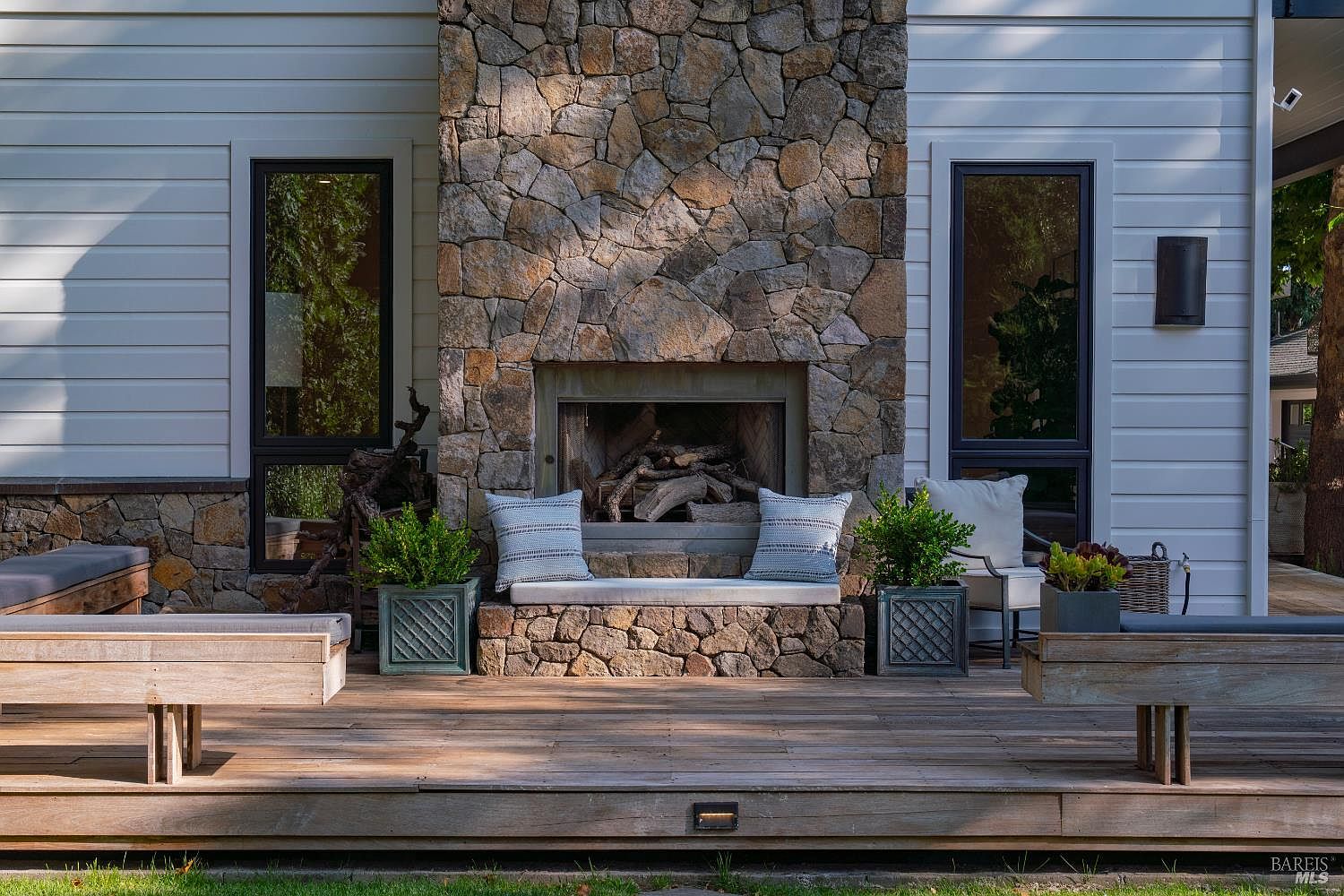 This inviting outdoor living space features a large stone fireplace as the centerpiece, flanked by two tall, modern windows and built-in stone seating with plush cushions. The expansive wooden deck is furnished with additional seating, including wooden benches and a chair, creating a perfect area for relaxation or entertaining. The clean lines of the white siding contrast beautifully with the rustic stone masonry, offering a sophisticated and cozy atmosphere.