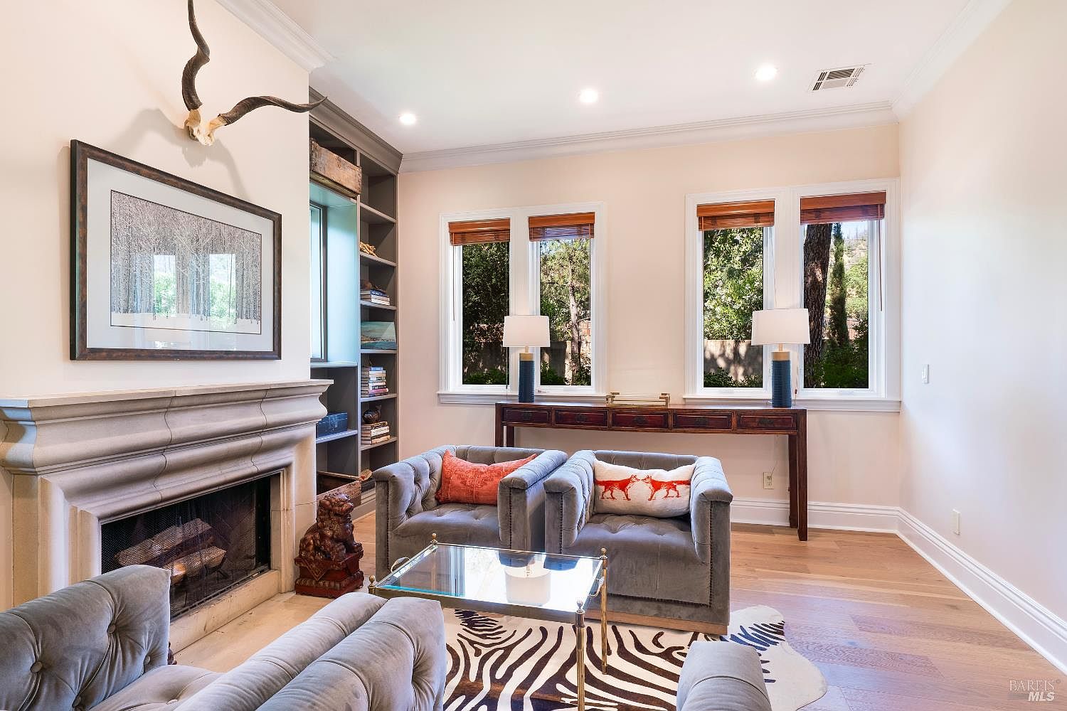 This elegant living room features a sophisticated fireplace with a stone mantel, topped by a large framed print and a decorative animal skull. The space is furnished with plush, tufted grey armchairs, a glass-topped coffee table, and a striking zebra-print rug, all set against light-toned walls and hardwood floors. Large windows with wooden blinds allow natural light to fill the room, while a built-in bookshelf and a dark wood console table add functional charm to the refined, cozy atmosphere.