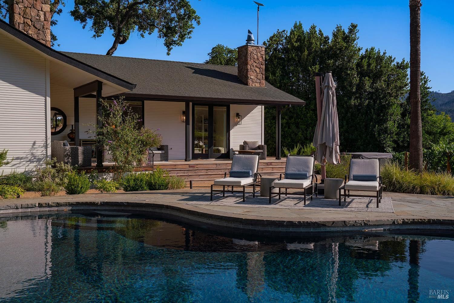 This inviting outdoor space features a dark-bottomed swimming pool in the foreground, leading to a spacious stone patio furnished with three lounge chairs and a large umbrella. The background showcases a charming single-story home with white siding, a stone chimney, and a covered wooden deck, all set against a backdrop of lush green trees and a clear blue sky. The scene captures a serene and luxurious backyard retreat, perfect for relaxation and entertaining.