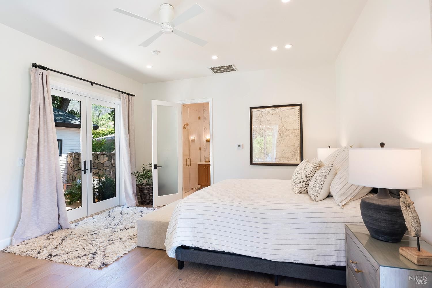 This bright and airy primary bedroom features a neutral color palette, light wood flooring, and a large bed dressed in striped linens. A set of French doors leads to an outdoor patio, while a frosted glass door provides access to an en-suite bathroom, creating a seamless flow between spaces. The room is accented by a framed map on the wall and a textured area rug, offering a serene and sophisticated atmosphere.