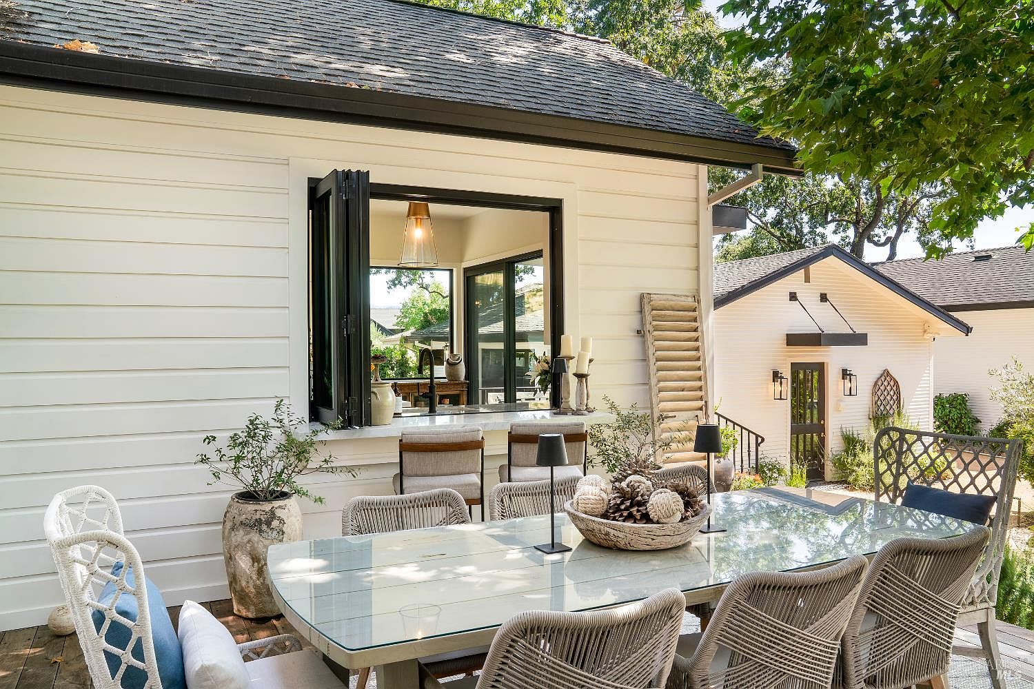 This inviting outdoor patio space features a large glass-topped dining table surrounded by woven chairs, perfect for al fresco entertaining. A unique pass-through window connects the indoor kitchen to the deck, creating a seamless flow between spaces, while the white horizontal siding and lush greenery provide a bright, coastal-inspired aesthetic. The scene is captured from a ground-level perspective, highlighting the functional connection between the home's interior and the serene backyard setting.