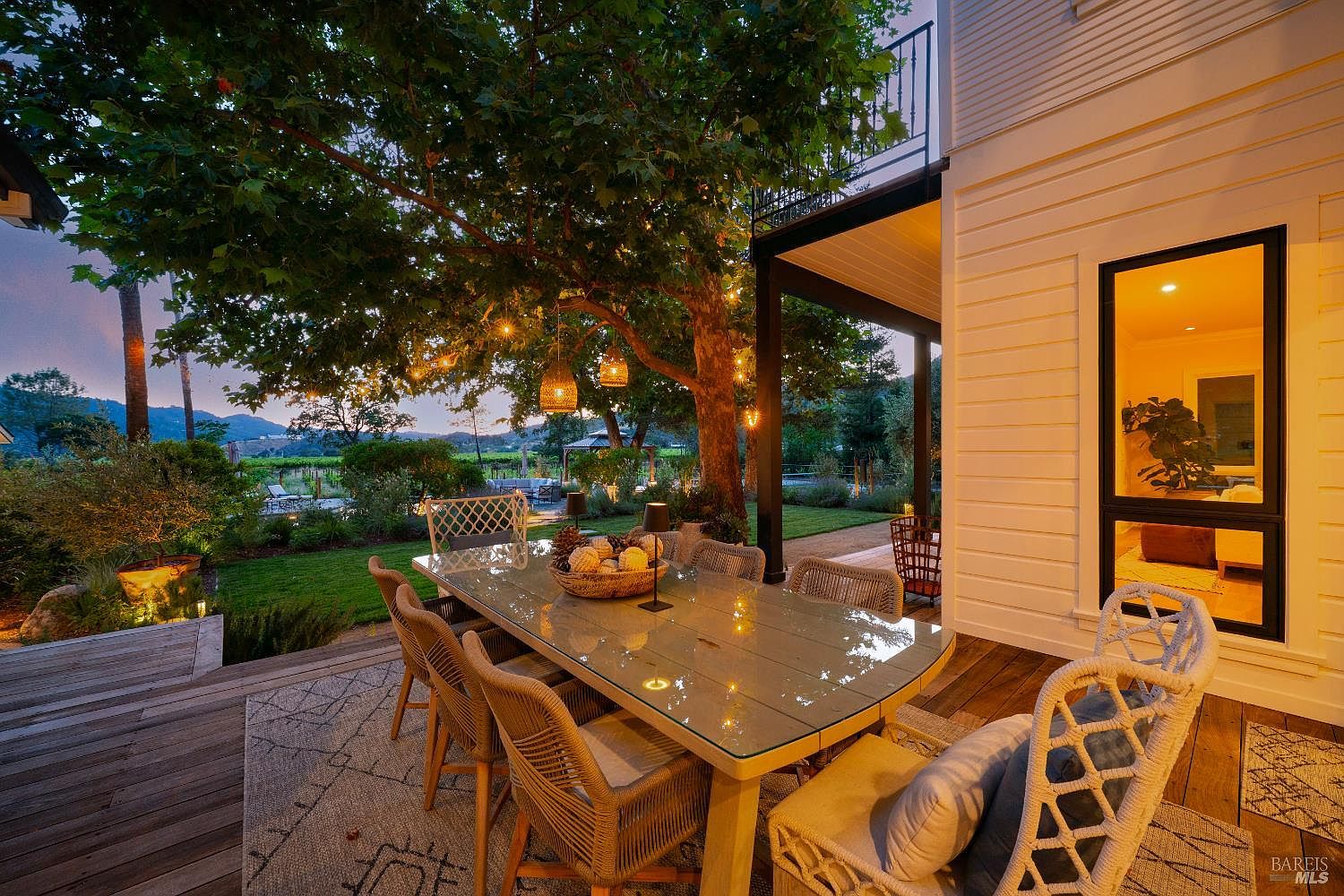 This inviting outdoor dining space features a glass-topped table surrounded by woven chairs, set upon a patterned rug on a wooden deck. The area is shaded by a large, mature tree adorned with warm string lights, creating a cozy atmosphere that overlooks a lush backyard and vineyard at dusk. The perspective captures the seamless transition between the indoor living space and the serene, well-manicured outdoor environment.