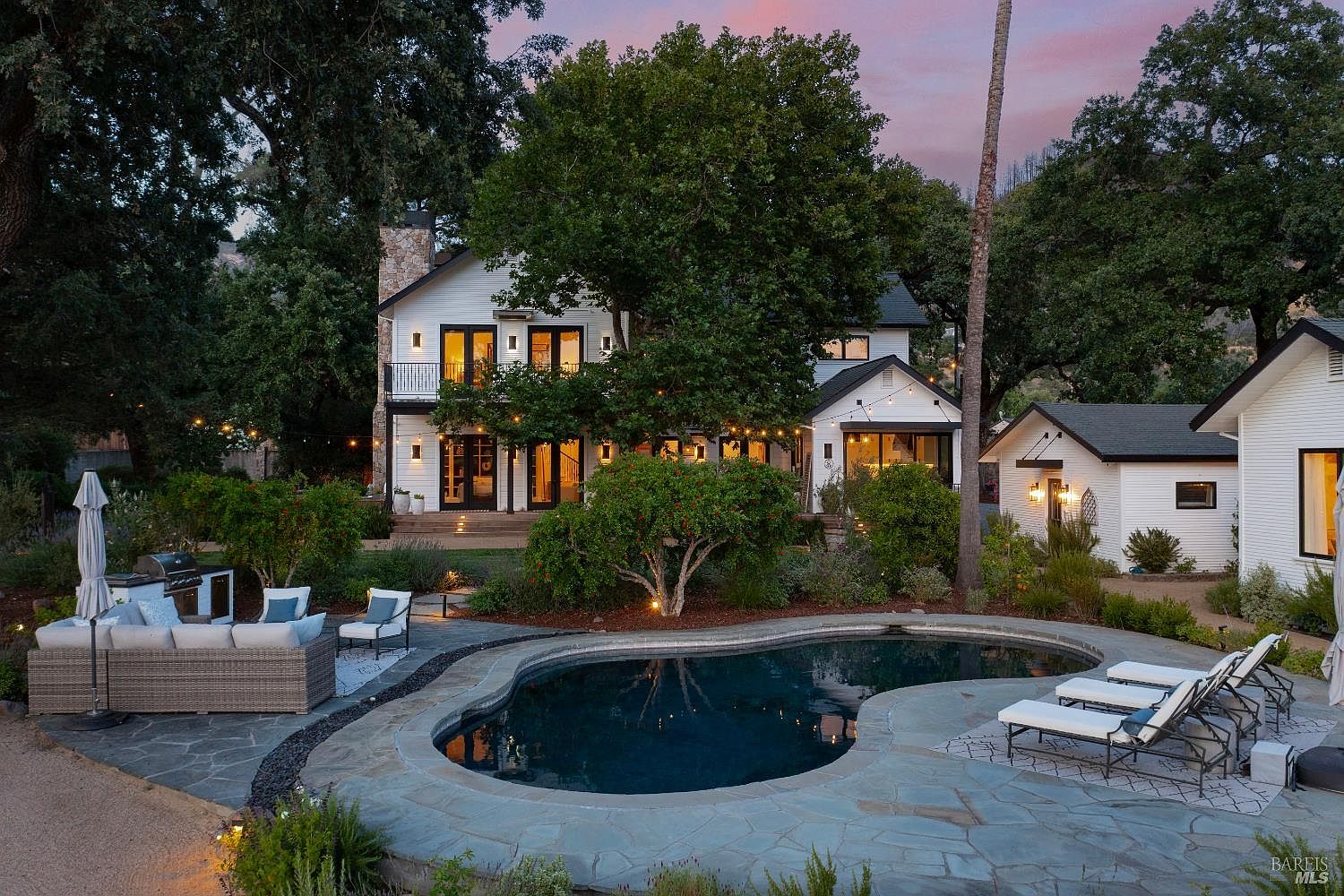 This stunning backyard scene features a custom-shaped swimming pool surrounded by natural stone decking, complemented by a comfortable outdoor lounge area and multiple chaise lounges. The white farmhouse-style home serves as a picturesque backdrop, illuminated by warm string lights and interior glow during the twilight hour. The lush landscaping and mature trees create a serene, private oasis perfect for relaxation and entertaining.