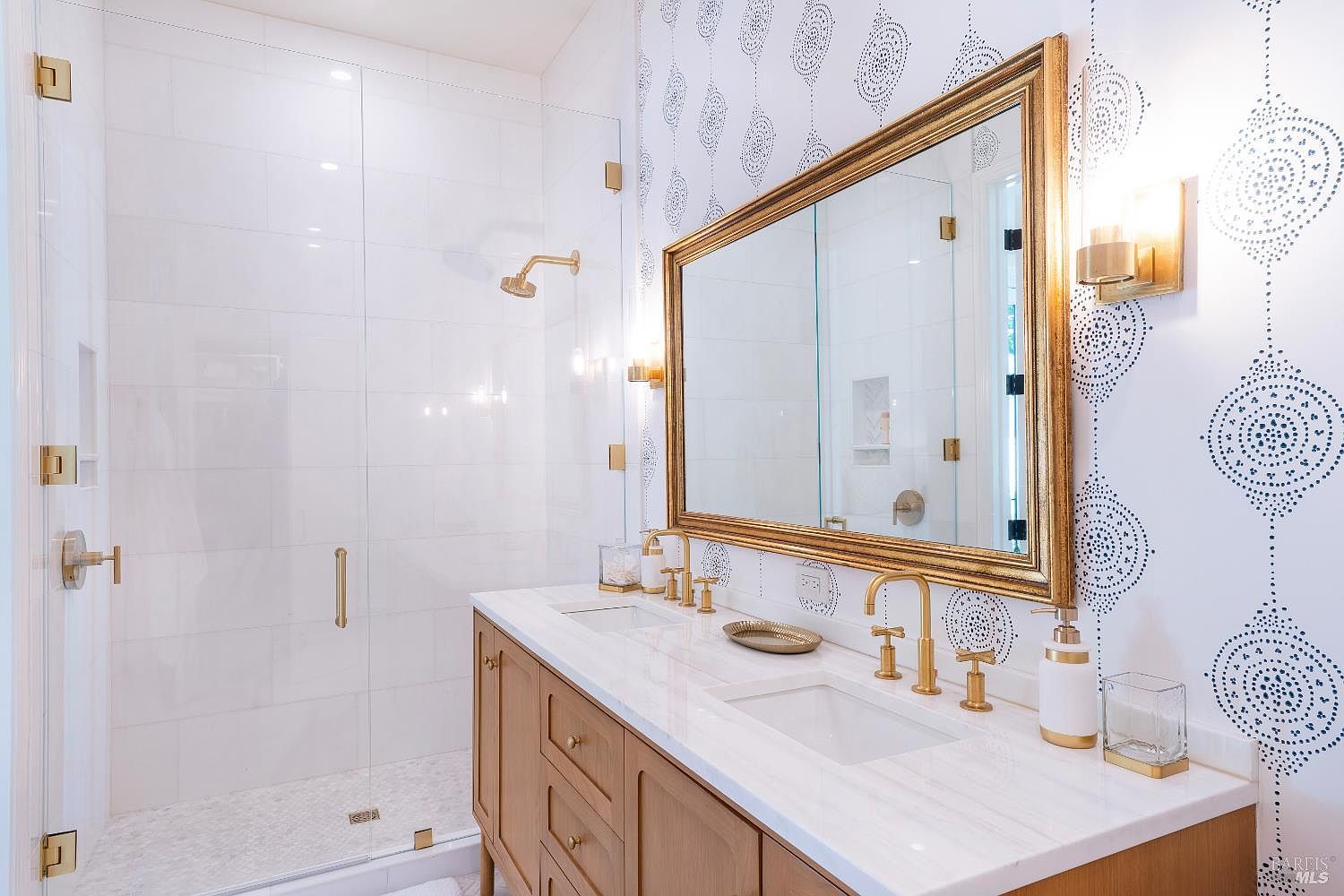 This elegant primary bathroom features a double vanity with a white marble countertop, warm wood cabinetry, and brushed gold fixtures. A large, gold-framed mirror hangs above the vanity, reflecting the stylish patterned wallpaper and the glass-enclosed walk-in shower with white subway tiles. The space exudes a sophisticated and modern aesthetic, blending clean lines with luxurious metallic accents.