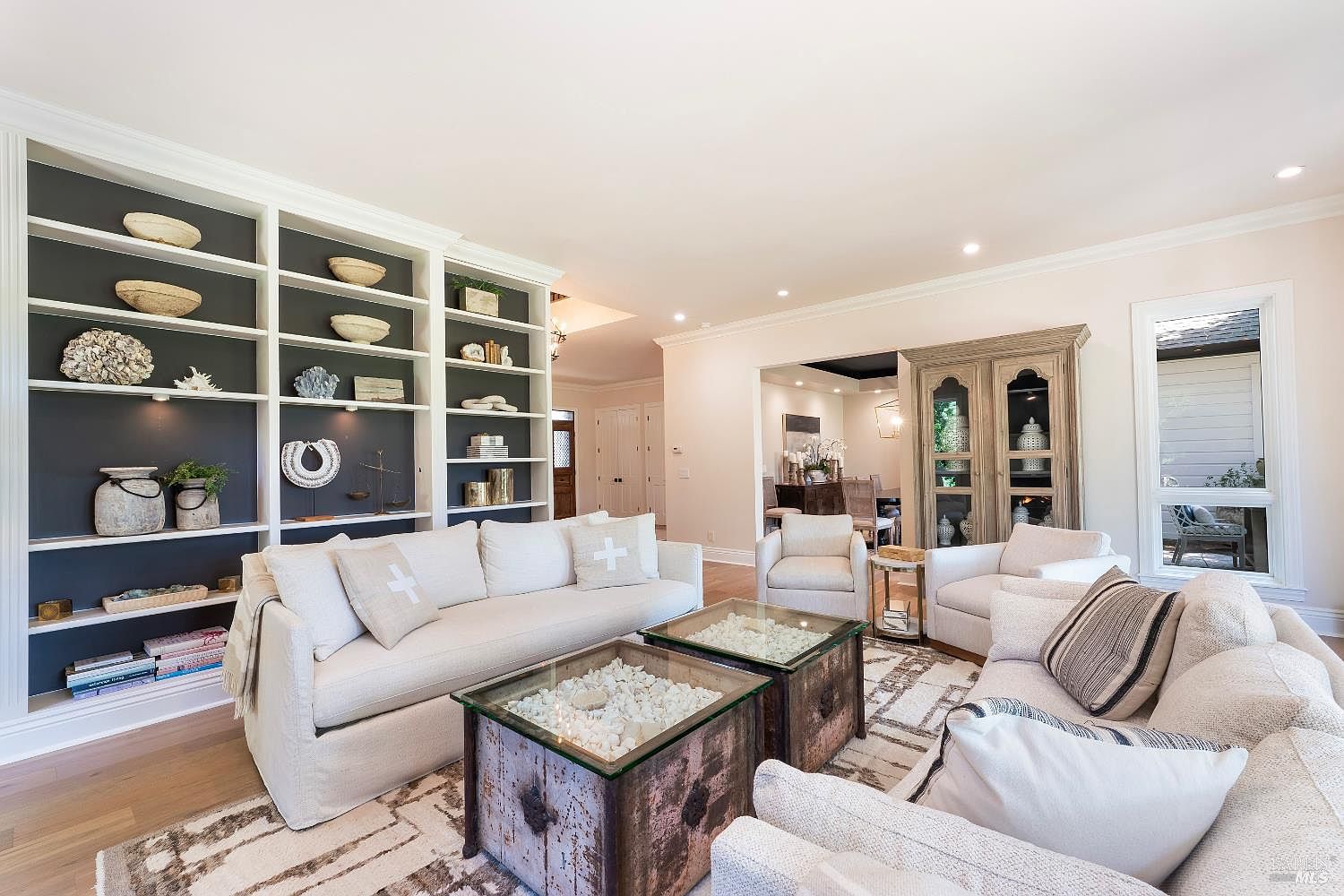 This elegant living room features a sophisticated neutral color palette, anchored by a large, custom built-in bookshelf with a dark charcoal backdrop. The space is furnished with plush, light-toned sofas and two unique, glass-topped coffee tables filled with decorative stones, creating a refined and inviting atmosphere. The open-concept layout flows seamlessly into the adjacent dining area, highlighted by recessed lighting and a classic wooden hutch.